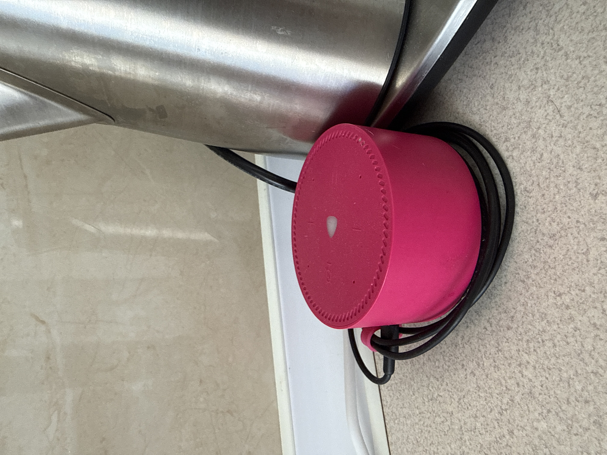 A close-up shot shows a vibrant pink cylindrical smart speaker resting on a speckled countertop. A black power cord is coiled around the base of the speaker, with a portion of the cord extending to the left. Behind the speaker, a brushed metal kettle with a black handle is visible, positioned on the right side of the frame. The background features a beige tiled wall and a white baseboard. The scene appears to be an indoor domestic setting, potentially a kitchen, located in Chișinău, Moldova.