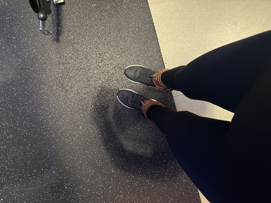 A person wearing black leggings and black sneakers with brown ankle straps stands on a speckled dark grey floor. A small portion of a light beige floor is visible in the upper right corner. To the left, a black metal arm from gym equipment is partially visible. The camera angle is overhead, capturing the individual from the knees down. The scene is illuminated with soft, diffused light.