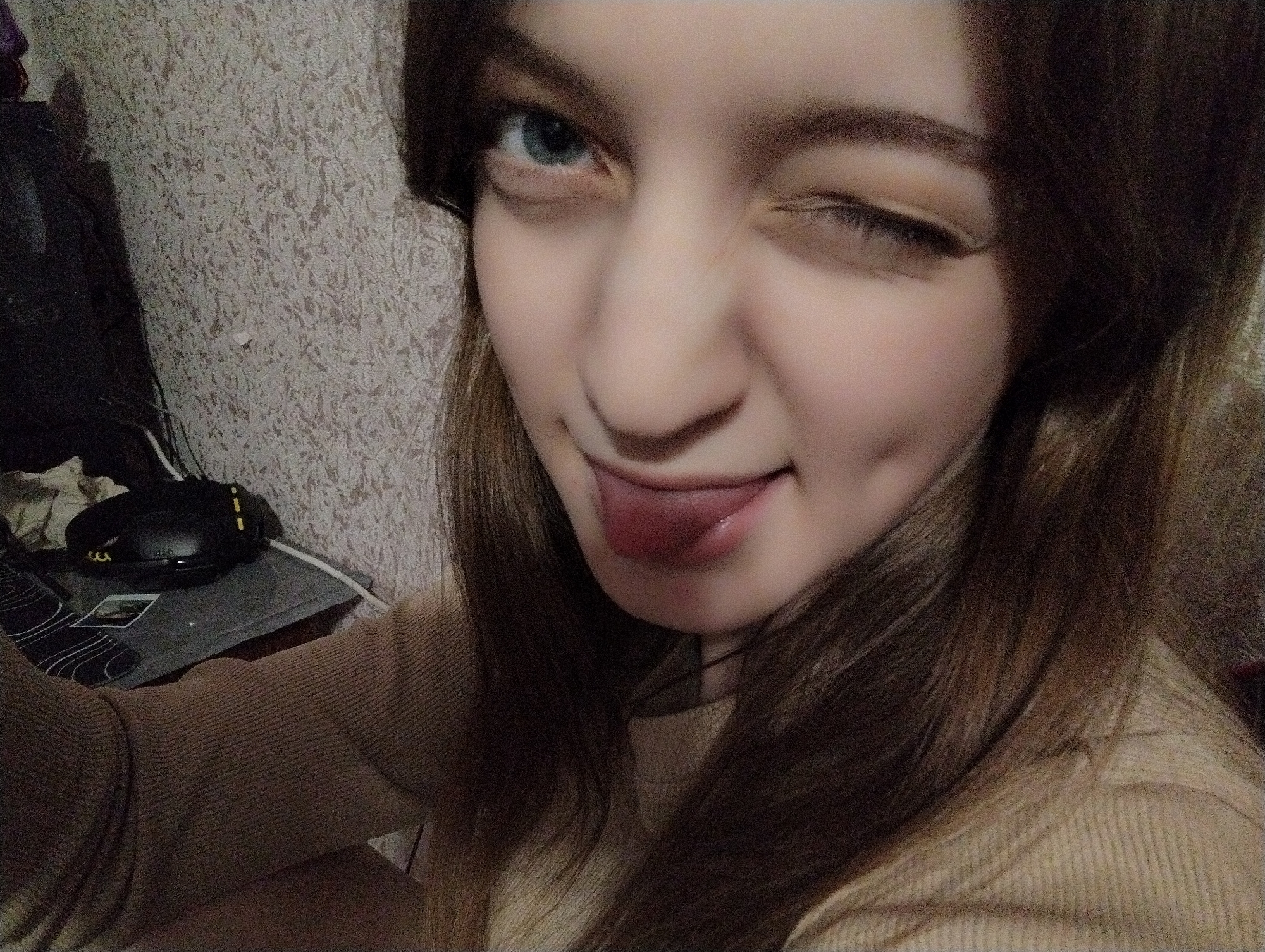 A young woman with light brown hair is seen in a close-up, selfie-style shot. She has fair skin, one eye open and blue, and the other is winked shut. Her tongue is playfully sticking out. She has a dimple on the right side of her face. She is wearing a light brown sweater. The background includes a textured wall and, to the left, a desk with a black computer mouse and other items. The location is Unknown, Unknown.