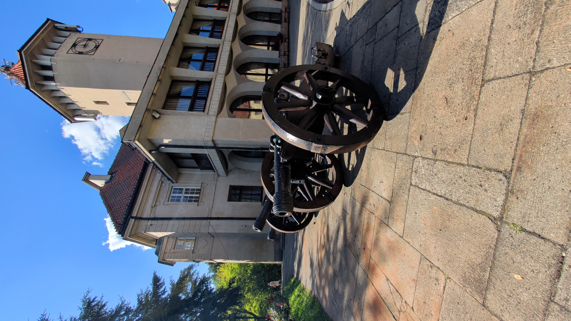 The image shows a sunny day in front of a building in Pułtusk, Poland. In the foreground, three antique cannons with wooden wheels are positioned on a stone-paved walkway. The building is made of light-colored stucco with a red-tiled roof and features a tall clock tower with a large, round clock face. The ground is paved with rectangular stone slabs, and there is a grassy area to the left with a few trees and bushes. A person in a light-colored outfit is walking on the grass in the background, and benches are visible near the building's arched entrance. The sky is blue with scattered white clouds.