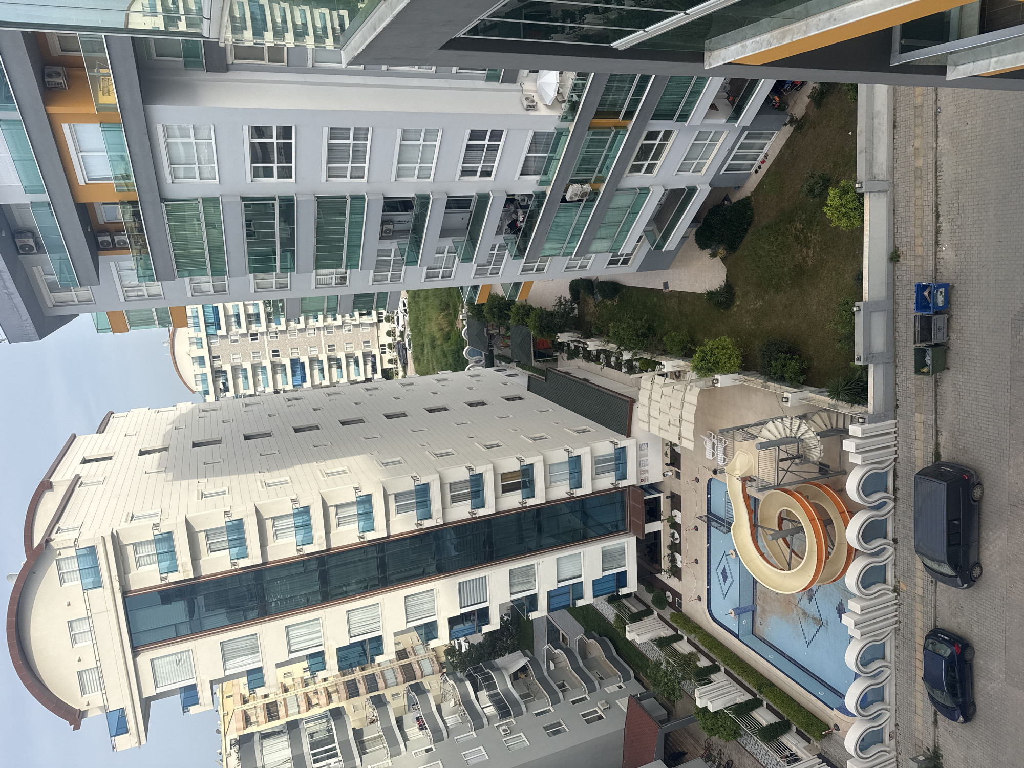 The image shows an aerial view of an urban residential area in an unknown city. Multiple high-rise apartment buildings are visible, featuring a mix of white, grey, and orange facades with glass balconies and windows. A swimming pool complex, including a water slide, is nestled between the buildings, alongside a small landscaped garden with trees and grass. Several parked cars are observed next to the paved sidewalk adjacent to the pool area. The sky is overcast, suggesting either a cloudy or hazy day. No people are clearly visible in the scene.