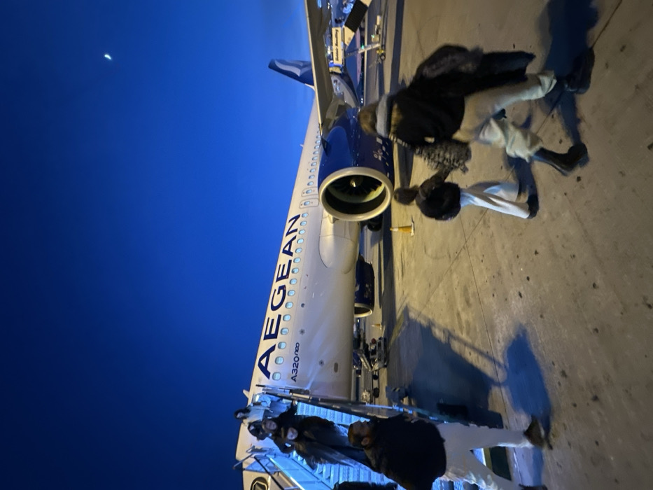 An Aegean A320neo aircraft is parked on the tarmac under a twilight sky. Several passengers are disembarking from a mobile stairway. The engine cowling of the plane is prominently displayed, along with the Aegean logo and "A320neo" printed on the fuselage. A crescent moon is visible in the upper left corner of the image. Some ground support equipment can be seen next to the plane. The photo appears to have been taken from an angle, possibly by someone on the ground.