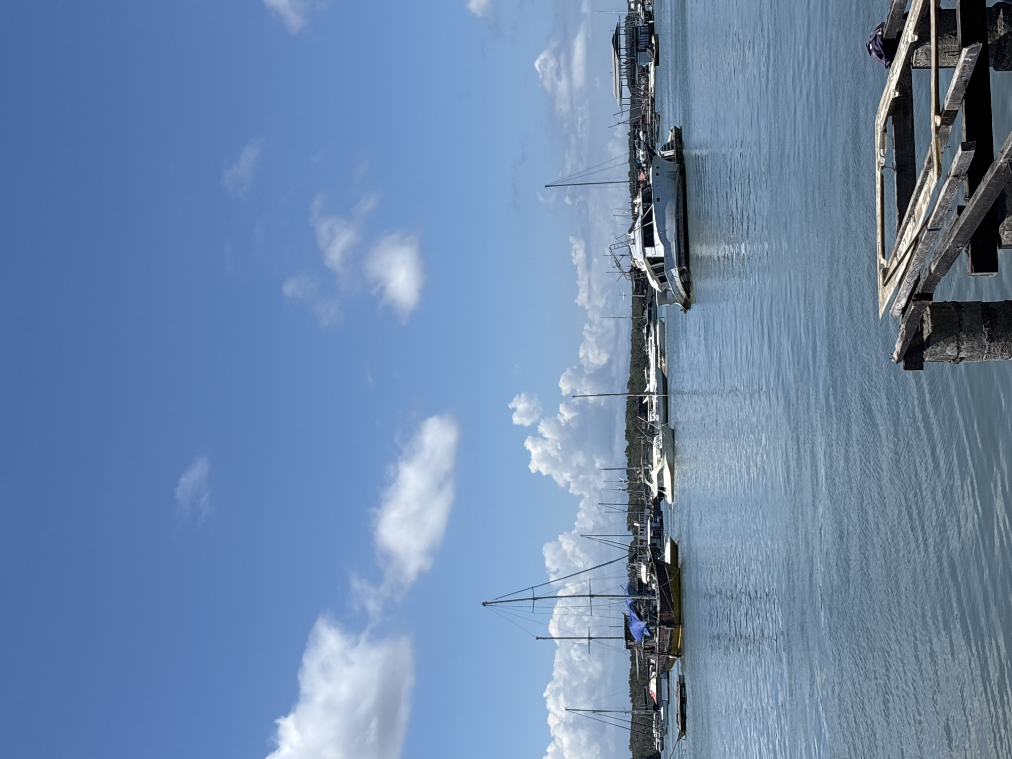 The image shows a harbor or marina under a partly cloudy, bright blue sky. Numerous boats, including sailboats and motorboats, are docked. There is a wooden structure or pier on the right. The location is likely in Denpasar, Indonesia.