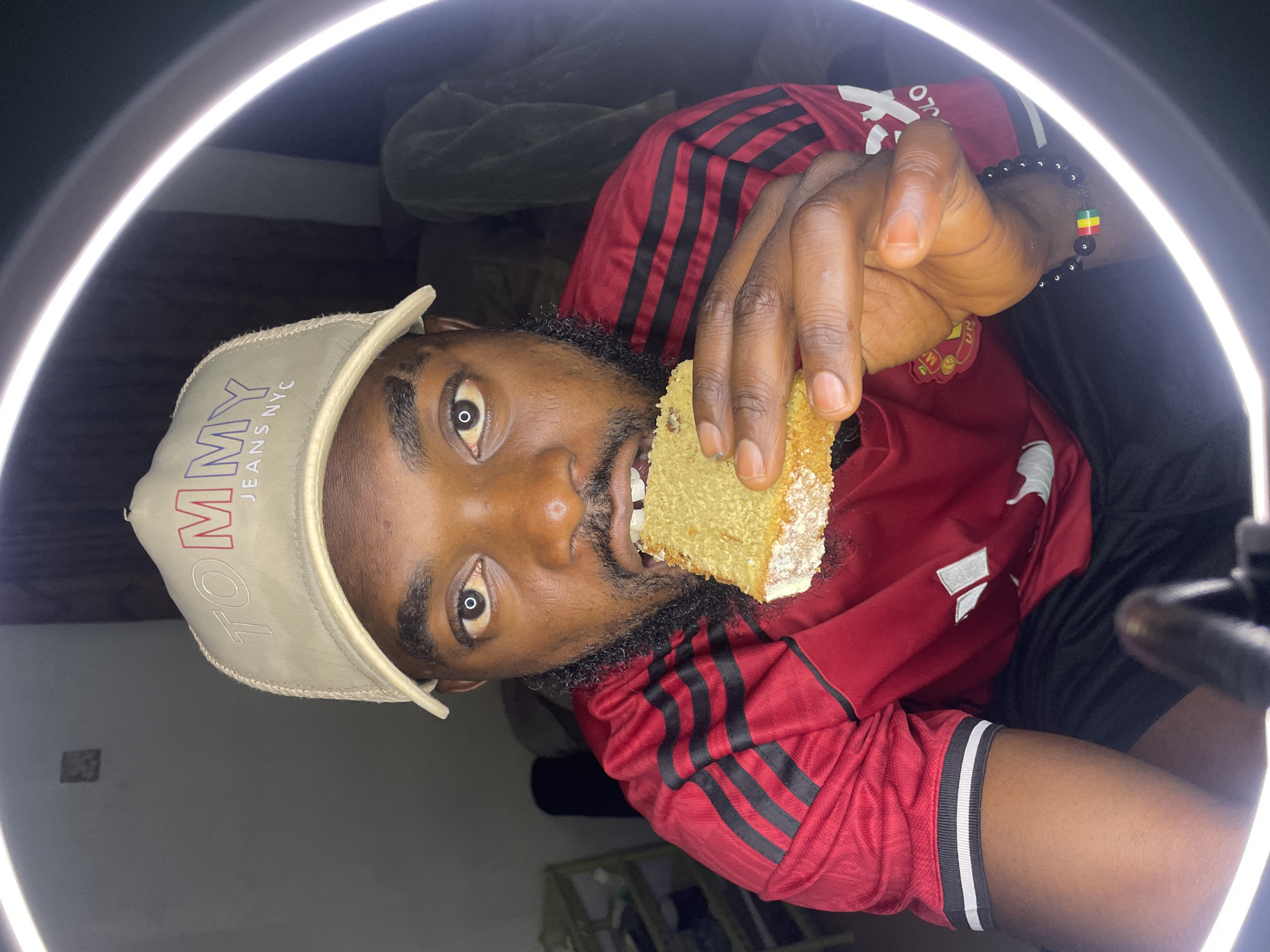 A dark-skinned man wearing a red and black Adidas shirt and a tan baseball cap with "TOMMY JEANS NYC" embroidered on it is eating a slice of cake. He is sitting in what appears to be an indoor setting, possibly a home, with a ring light illuminating his face. The background is dimly lit and shows a blurred shelving unit on the left and a draped cloth on the right. A beaded bracelet with red, yellow, and green beads is visible on his wrist. The man is looking directly at the camera with a neutral expression. The image does not contain any information or visual cues that suggest a specific location in the city of Jos, Nigeria.
