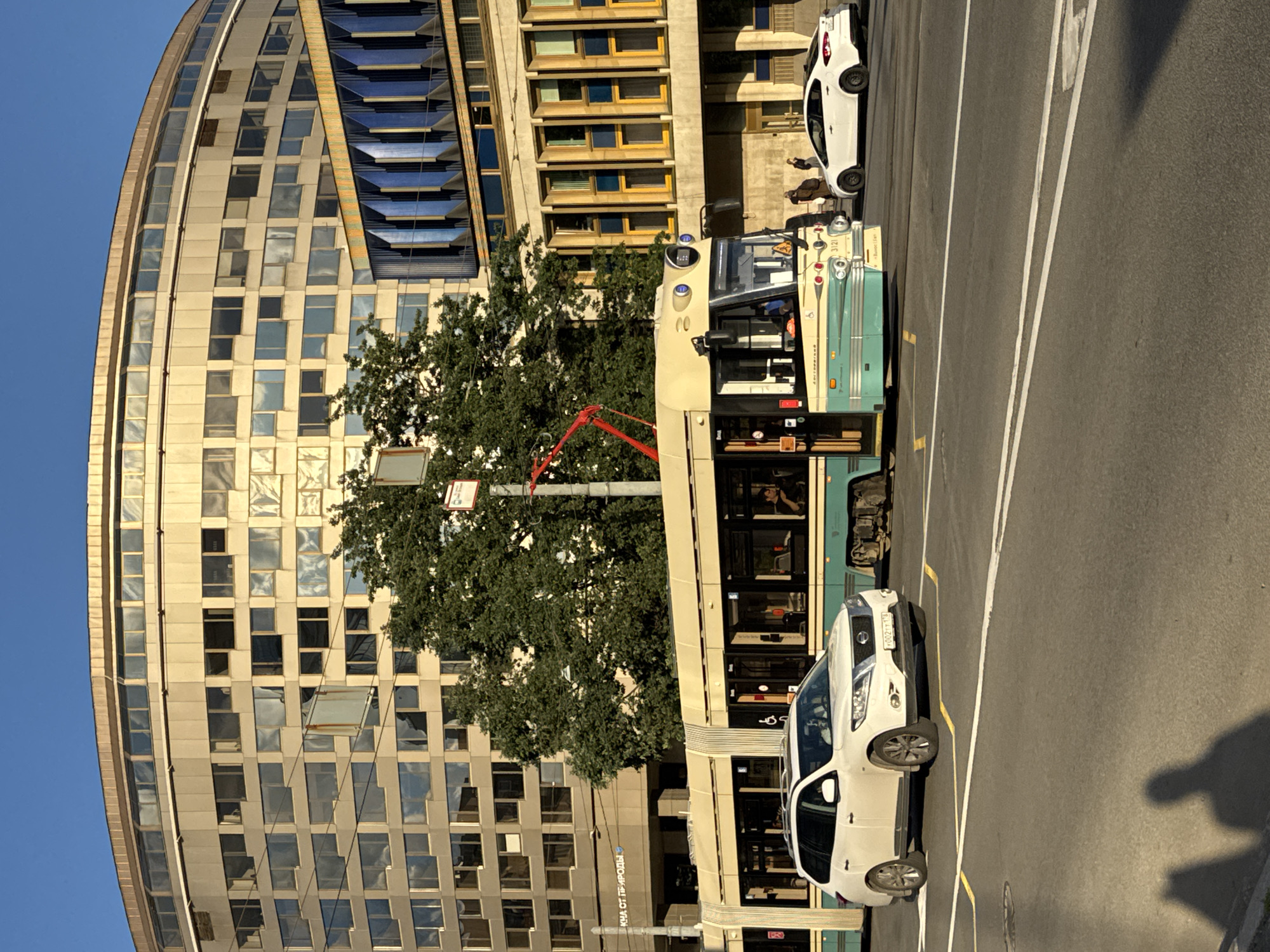 In an urban environment, a white SUV is parked on a street, facing left. Behind the SUV, a cream and teal tram travels forward. A white sedan is visible to the right of the tram. The background features a large, circular beige building with numerous windows, and to its right, a blue and yellow building with a patterned facade. Trees are present between the buildings. The sky is clear and blue. Shadows of two people are cast on the road in the foreground.