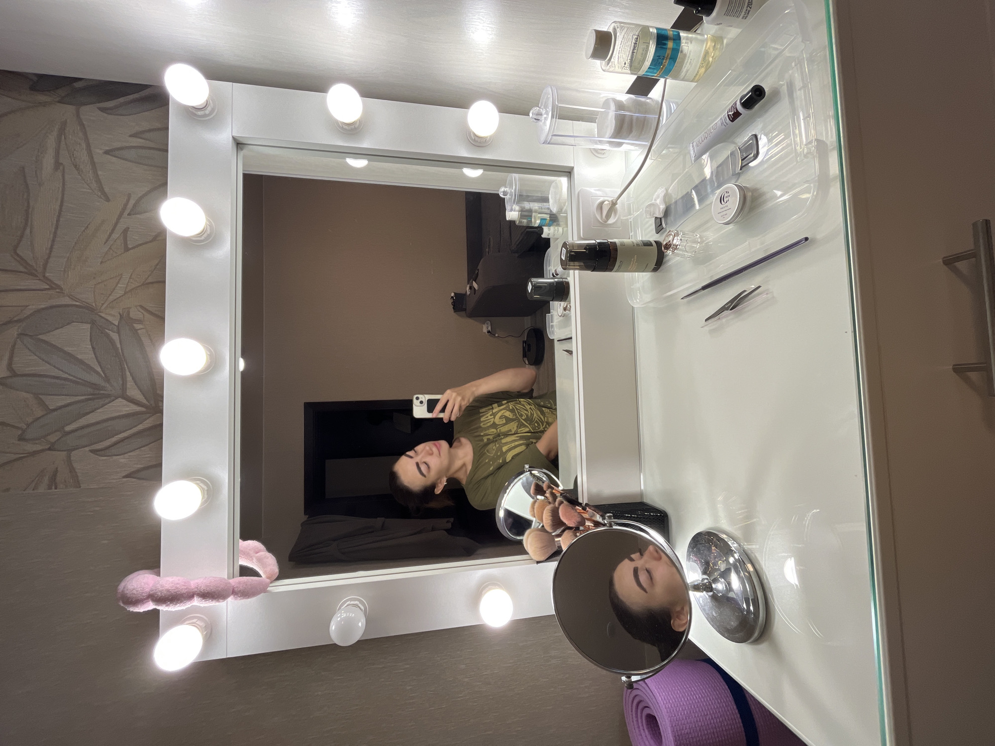 The image depicts an indoor scene featuring a vanity table with a large, illuminated mirror. A woman is visible in the mirror, taking a selfie with her phone. On the vanity, various makeup brushes are arranged in a holder, along with several cosmetic products and accessories. A pink fluffy headband is draped over the top of the mirror. To the left of the vanity, a rolled-up purple yoga mat is visible. The room appears to be a personal dressing area or a makeup studio. There is no information in the image to suggest the location is in Chaikovsky, Russia.