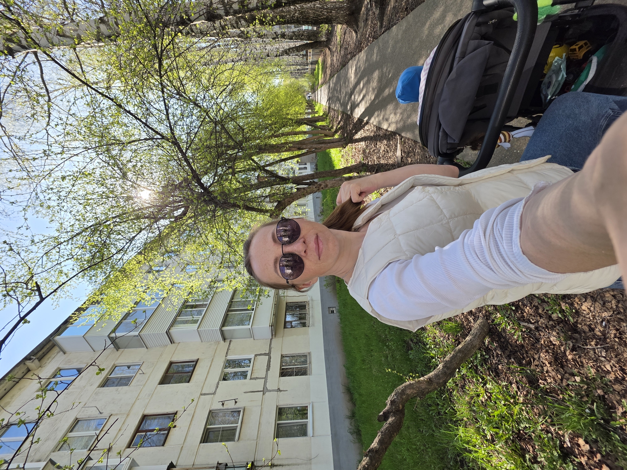 A selfie of a woman wearing sunglasses, a white long-sleeve shirt, and a white vest. She is positioned next to a baby stroller. The location appears to be in the city of Chaikovsky, Russia, indicated by the architectural style of the building to the left of the woman and the prevalence of birch trees along the sidewalk. The background features a paved walkway lined with mature trees, a building with multiple windows, and patches of grass.