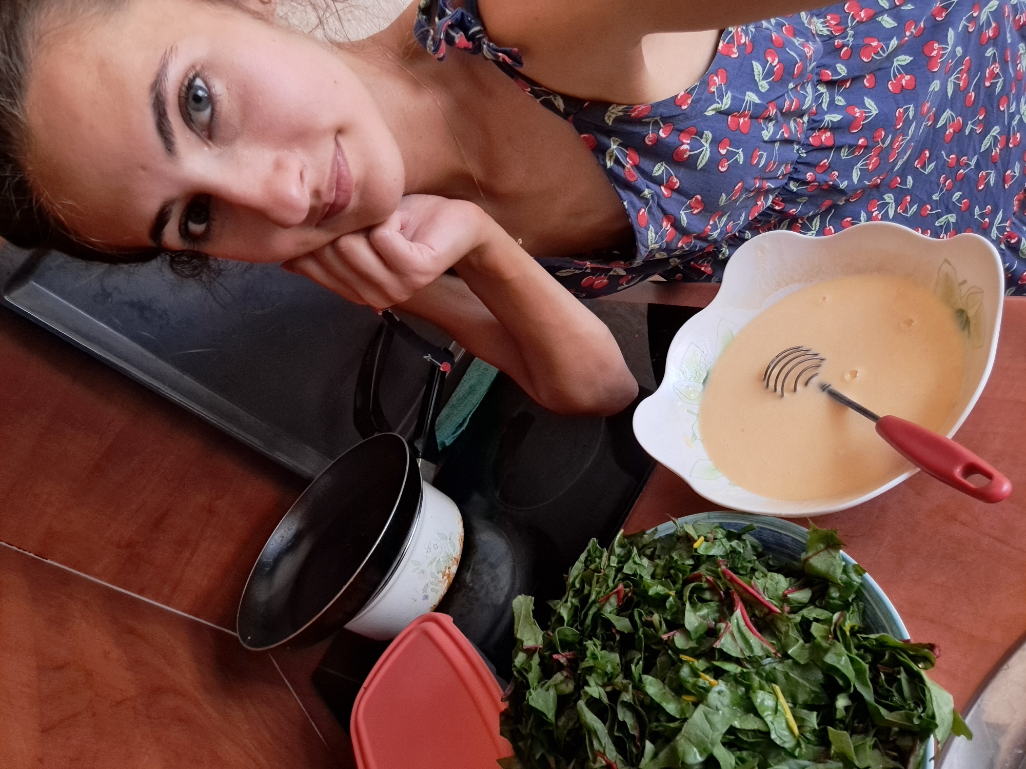 A young woman with dark hair and blue eyes is in a kitchen, taking a selfie. She is wearing a blue dress with a cherry print. In front of her are a bowl of chopped leafy greens, a bowl of yellow batter with a whisk, and a pot on a stovetop. The kitchen has an orange tiled backsplash and a dark countertop.