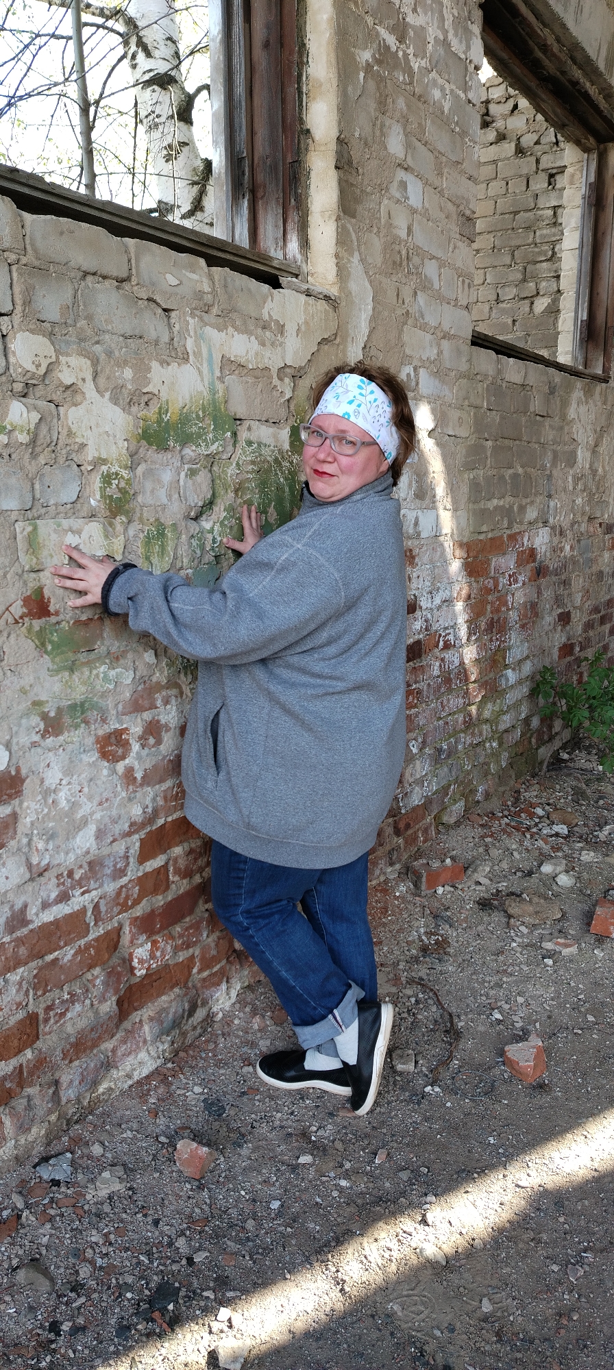 A woman stands near an old brick wall in what appears to be an abandoned building or ruin in Зипуново, Russia. The woman, wearing glasses, a white and blue floral headband, a gray sweatshirt, jeans, and black shoes, places her hands on the dilapidated wall. The wall is made of both red and white bricks, with patches of exposed plaster and some green moss or paint. Debris and rubble litter the ground around her feet. Light streams in from open window frames in the background.