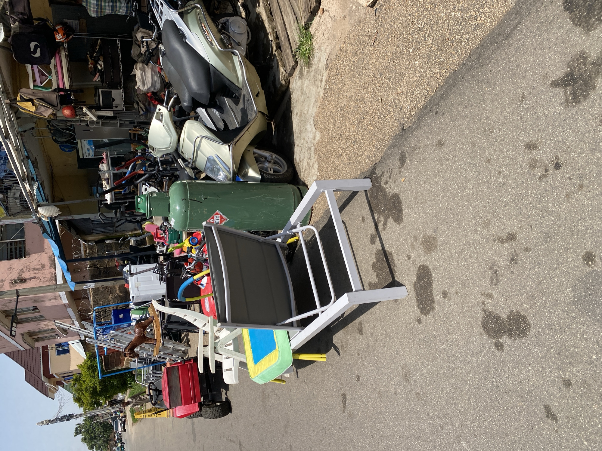 In a street scene in Jos, Nigeria, a variety of items are displayed for sale or storage along the side of a paved road. In the foreground, a white lounge chair with a colorful cushioned seat, featuring yellow legs, sits on the asphalt. Behind it, a large green gas cylinder is positioned next to a light-colored scooter. Further back, a red riding lawn mower is visible, along with a brown rocking horse on a display rack. Other objects scattered in the area include a washing machine, a blue cooler, and various metal items. A man in a plaid shirt stands on the right side of the frame, observing the scene. The environment appears to be an outdoor market or a storage area next to a building with a pink facade. The road surface is asphalt with visible water stains.