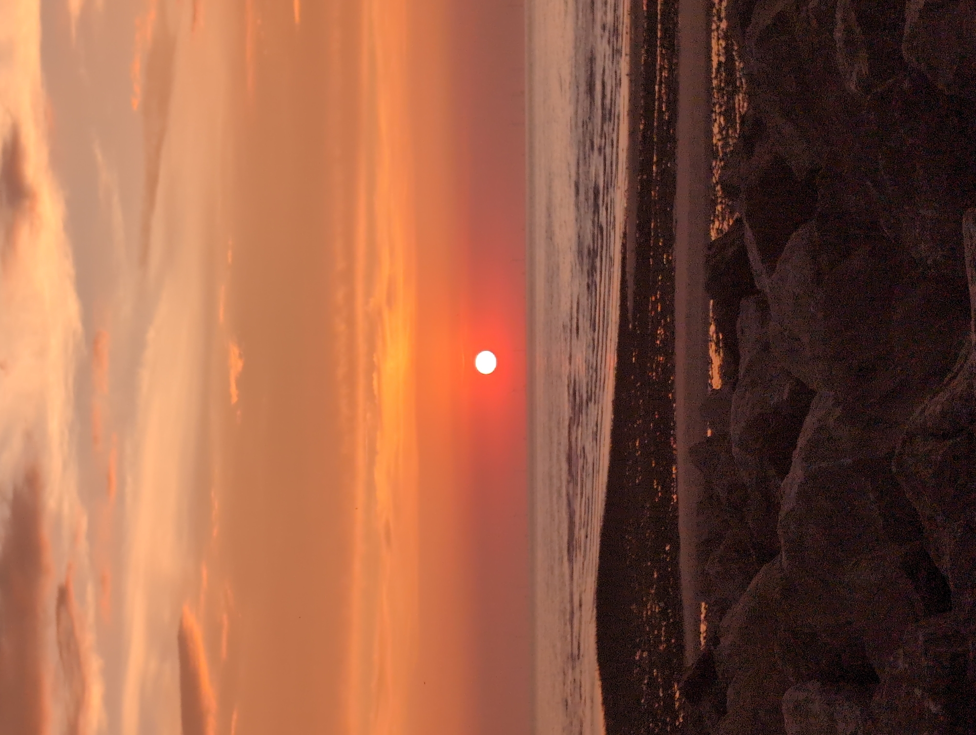 The image depicts a sunset over the sea, viewed from the beach in Le Tréport, France. The sky is filled with warm hues of orange and pink, with scattered clouds. The sun, a bright orb, is low on the horizon, casting a warm glow over the water. The sea is calm, with gentle waves lapping at the shore. The foreground of the image is dominated by dark, rocky terrain, suggesting a breakwater or natural rock formations along the coastline. The overall scene conveys a sense of tranquility and natural beauty.