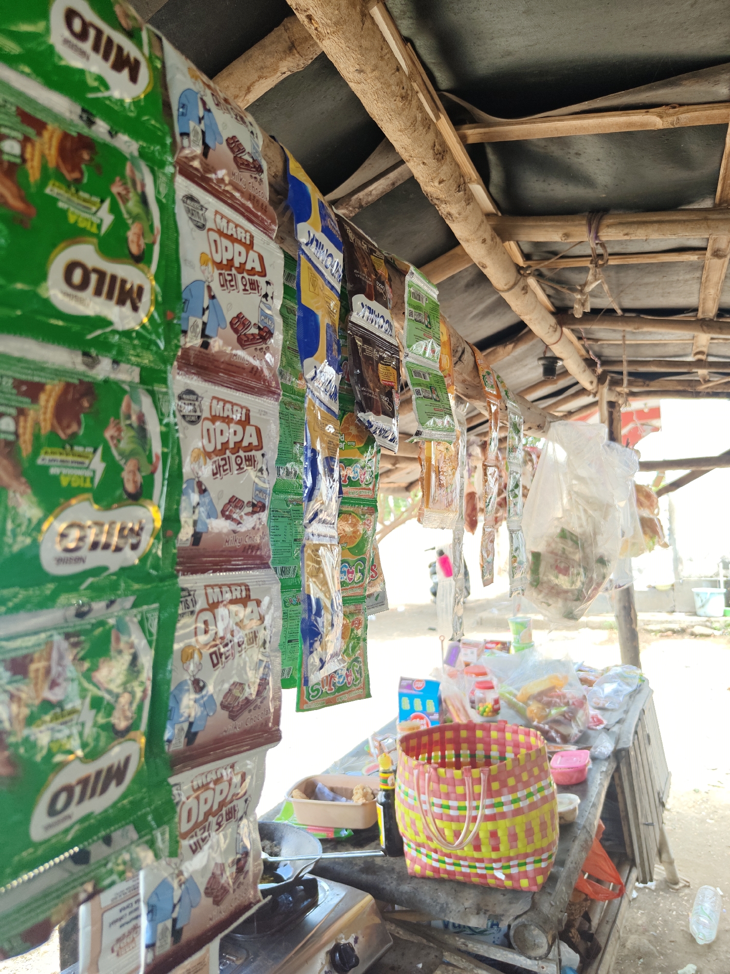 The image depicts an outdoor stall, likely a small convenience store or roadside vendor, in Cermee, Indonesia. The stall has a rustic construction, with a wooden frame and a dark tarp for a roof. Various food and drink packets are displayed, hanging from the roof structure. Prominently visible are green packets of "Milo" and several packets of "Mari Oppa" with Korean text and images of chocolate bars. Other items include small packaged snacks, candies, and some plastic bags filled with goods. On a table beneath the hanging items, there is a colorful woven basket, some containers, and a small stove. The ground appears to be dirt. In the background, a blurry image of what might be a motorcycle and some buildings can be seen, suggesting a semi-urban or rural setting. The lighting indicates it is daytime.