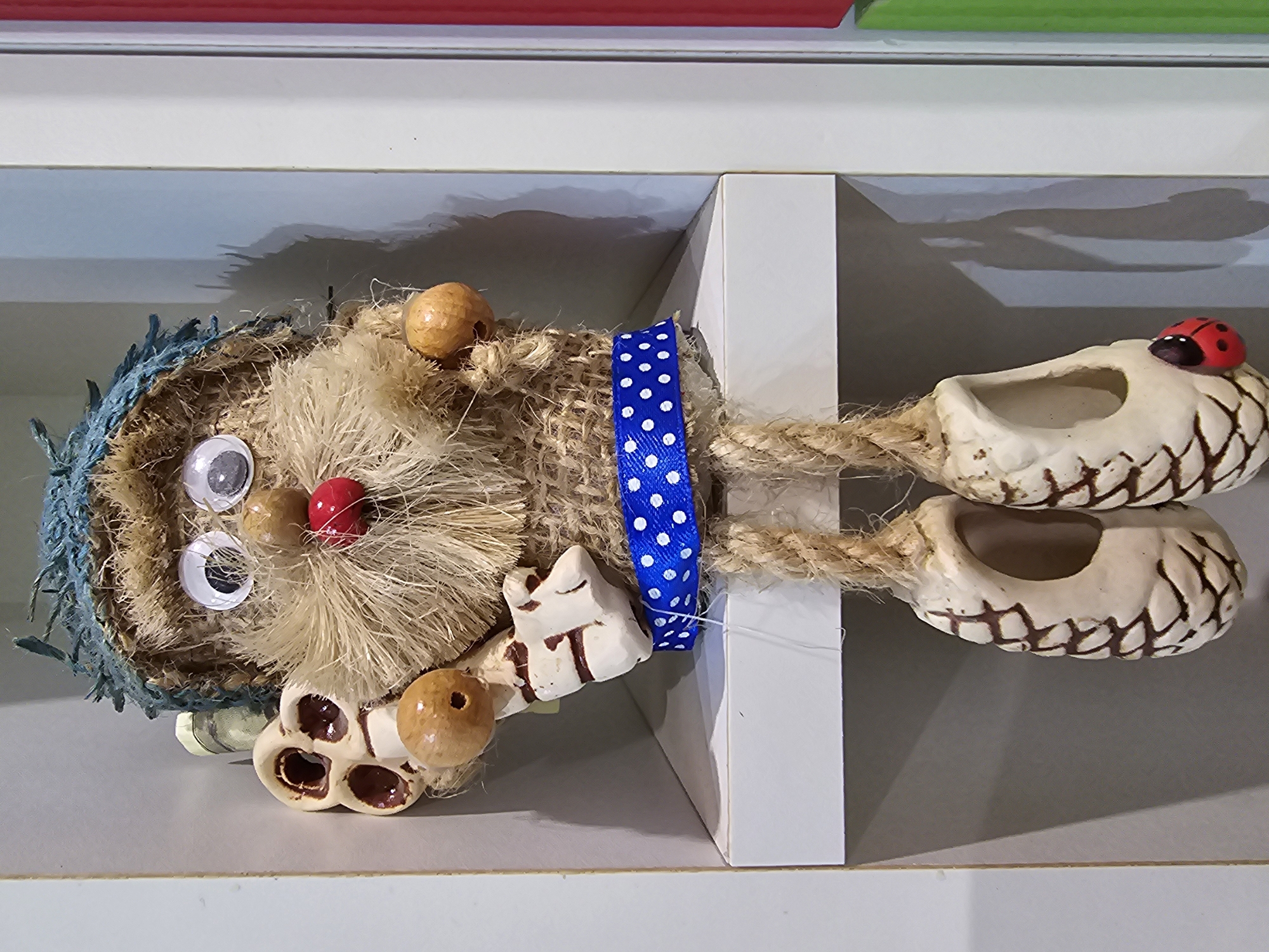 A stuffed doll, approximately 2 feet tall, is sitting on a white shelf. The doll has a rough, straw-like texture and is wearing a blue, braided hat. It has large googly eyes, a red bead for a nose, and wooden beads for cheeks and ears. The doll's body is wrapped in burlap, cinched with a blue and white polka dot ribbon. Its arms are made of twigs, and one arm is holding a beige, sculpted object with four round holes. The doll's legs are made of braided rope, and each leg ends in a hollow, beige shoe shaped like a pinecone, decorated with brown lines. One of the shoes has a small red ladybug charm attached. The shelf is part of a white, built-in cabinet, which is positioned in a corner of a room. The background is a neutral gray wall. The lighting suggests an indoor setting, possibly a home or a shop. This scene is located in the city of Kalinovka, Russia.