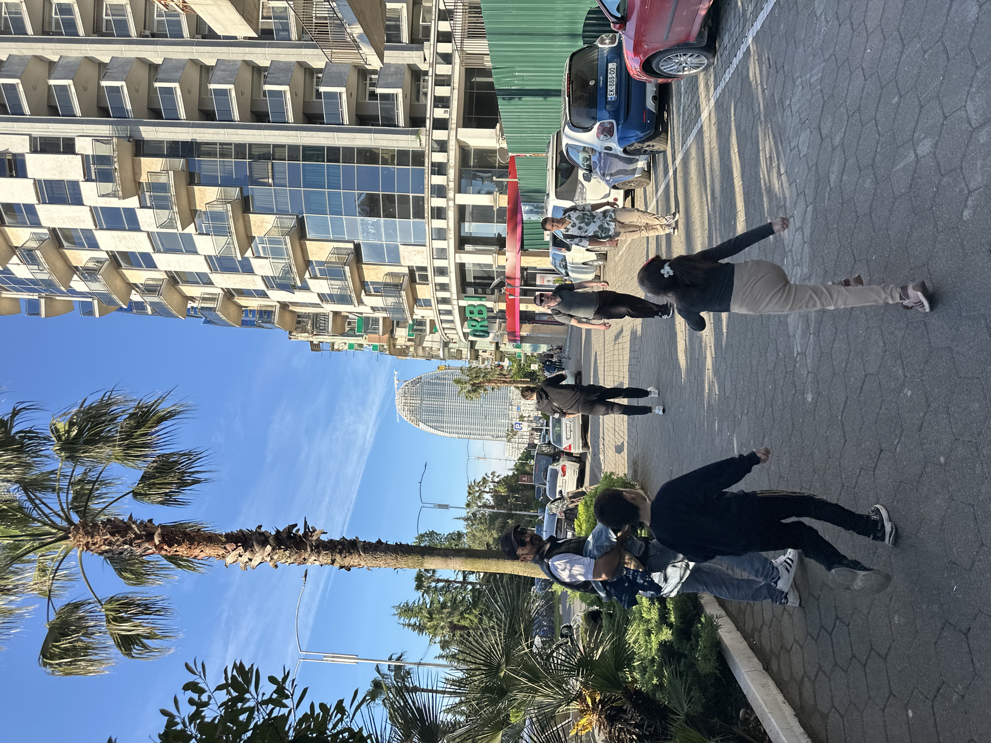 The image shows a street scene in Batumi, Georgia. In the background, there is a tall building with a checkered facade, as well as a tall, cylindrical, modern building that could be a hotel or a residential tower. Several parked cars are visible along the street. A group of people is present on the sidewalk, appearing to be standing or walking. A palm tree stands along the edge of the sidewalk, adding a tropical element to the urban environment. The sky is blue with some clouds. The photo appears to be taken during the daytime.