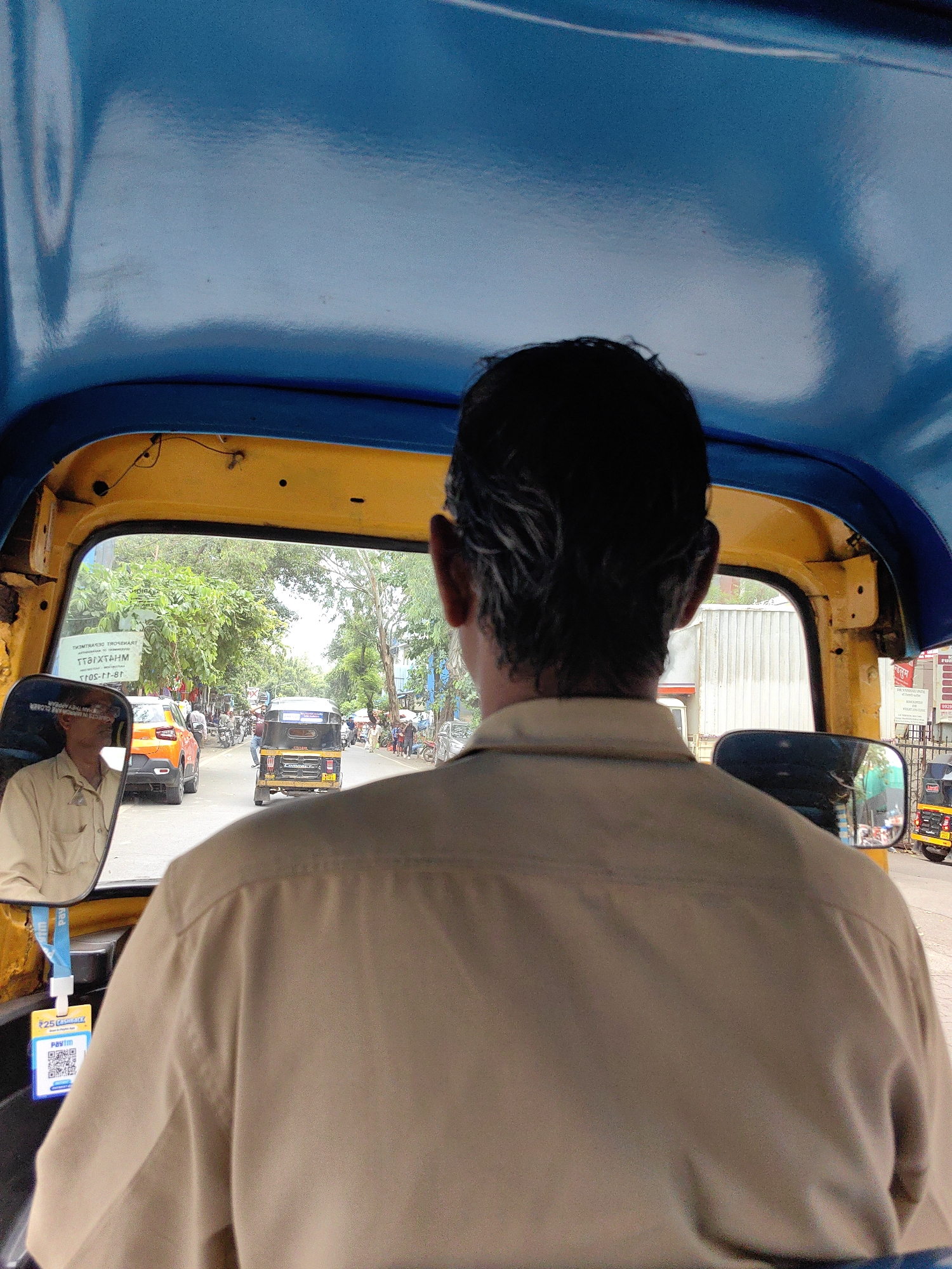 The image depicts the interior of an auto-rickshaw, looking forward through the windshield. The driver, a man with dark hair, is seen from behind, wearing a light brown shirt. The interior of the auto-rickshaw is bright yellow, with a blue canopy. A side mirror on the left reflects the driver's face and a sign with text in Marathi. Outside the windshield, a street scene is visible, with other vehicles, including another auto-rickshaw and a car, and pedestrians. Trees line the street, and buildings are present in the background. The image is likely taken in Mumbai, India, given the context of auto-rickshaws and the Marathi text on the sign. A Paytm QR code hangs from a lanyard inside the auto-rickshaw.