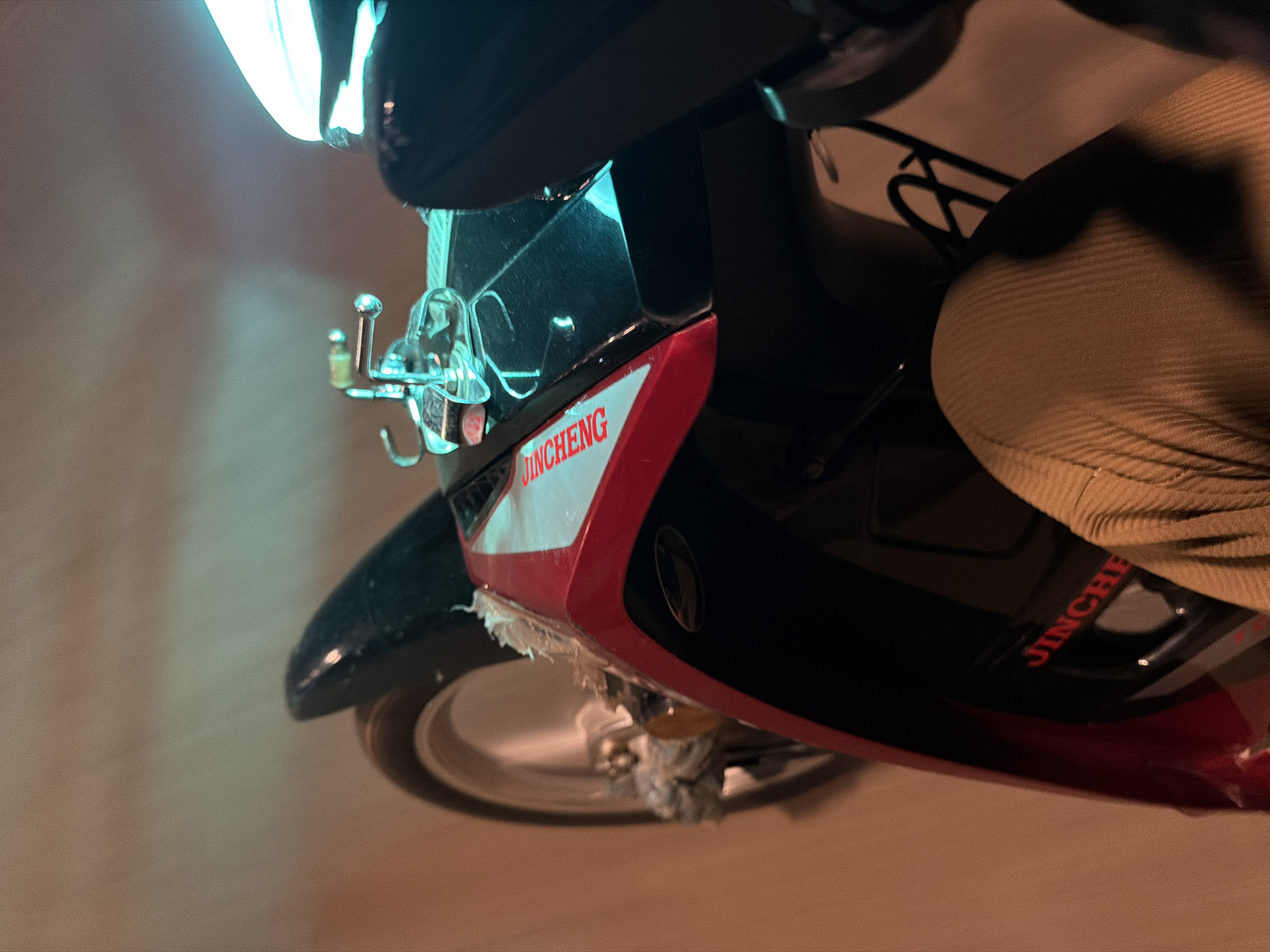 The image shows a close-up, eye-level, point-of-view shot from the perspective of someone riding a red and black motorcycle at night. The motorcycle's front wheel is partially visible on the left, with a fuzzy, light-colored material wrapped around the hub. The motorcycle's body is red and black, with a white stripe and red lettering that reads "JINCHENG" on the side. A chrome-colored fuel cap with a small hook is visible on the top of the motorcycle's body, reflecting a blue light. The rider's right leg, clad in tan corduroy pants, is prominent in the foreground, suggesting the motorcycle is in motion. The background is blurred, with streaks of orange and blue light, indicating movement through a dimly lit environment, possibly a street in Bukuru, Nigeria. The lighting suggests artificial light sources, such as streetlights or vehicle headlights, are present.