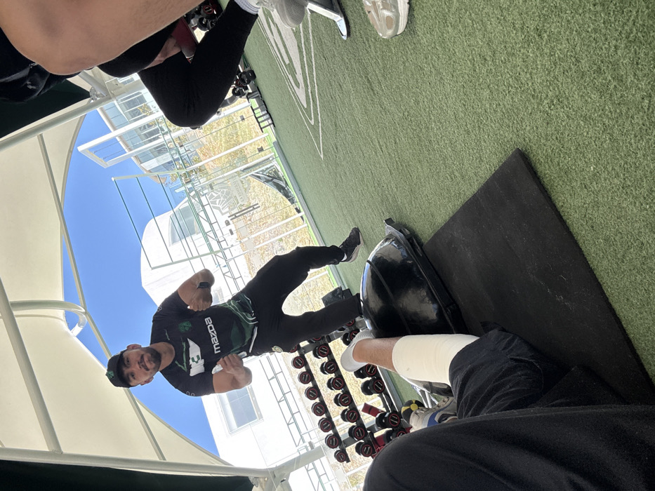 A man in a Mazda shirt and cap stands with clenched fists in a gym. Dumbbells are lined up near him. A person sits in the foreground with a wrapped leg and a blue and white shoe. Green turf covers the floor, and a black mat is visible near the seated person. Outside, a white building is visible under a blue sky. The scene is partly shaded by a white awning.