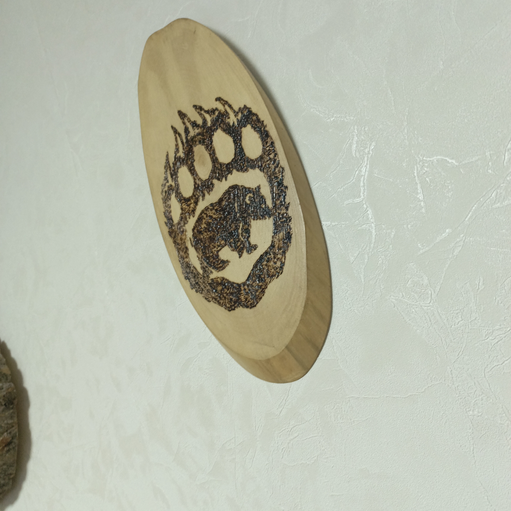 The image displays a light-colored, oval-shaped wooden plaque mounted on a textured wall. The plaque is angled, with its upper left side closer to the viewer, showcasing its depth.
The central feature of the plaque is an intricate pyrography (wood-burned) design. This design consists of a large bear's paw print, detailed with distinct claws and paw pads, encompassing a smaller, full-body depiction of a bear cub. The cub is rendered with visible fur texture, and its features, including its eyes and snout, are clearly delineated. The wood-burned lines are dark brown, creating a strong contrast against the pale, natural wood of the plaque.
The background is a textured wall, appearing in a light off-white or very light beige color. The texture is subtle and irregular, suggesting either a specific type of wallpaper or a textured paint finish. A small, partial glimpse of another object, resembling a natural piece of wood or stone with a rough surface, is visible on the far left edge of the frame, casting a soft shadow on the wall.
The scene is static, focused on the decorative wooden artwork. There are no people present, and no activity or event is taking place. The lighting suggests an indoor setting, possibly under artificial light, but the specific time of day is not evident. No visible text is present on the plaque or elsewhere in the image. The overall impression is one of rustic, nature-inspired decor.