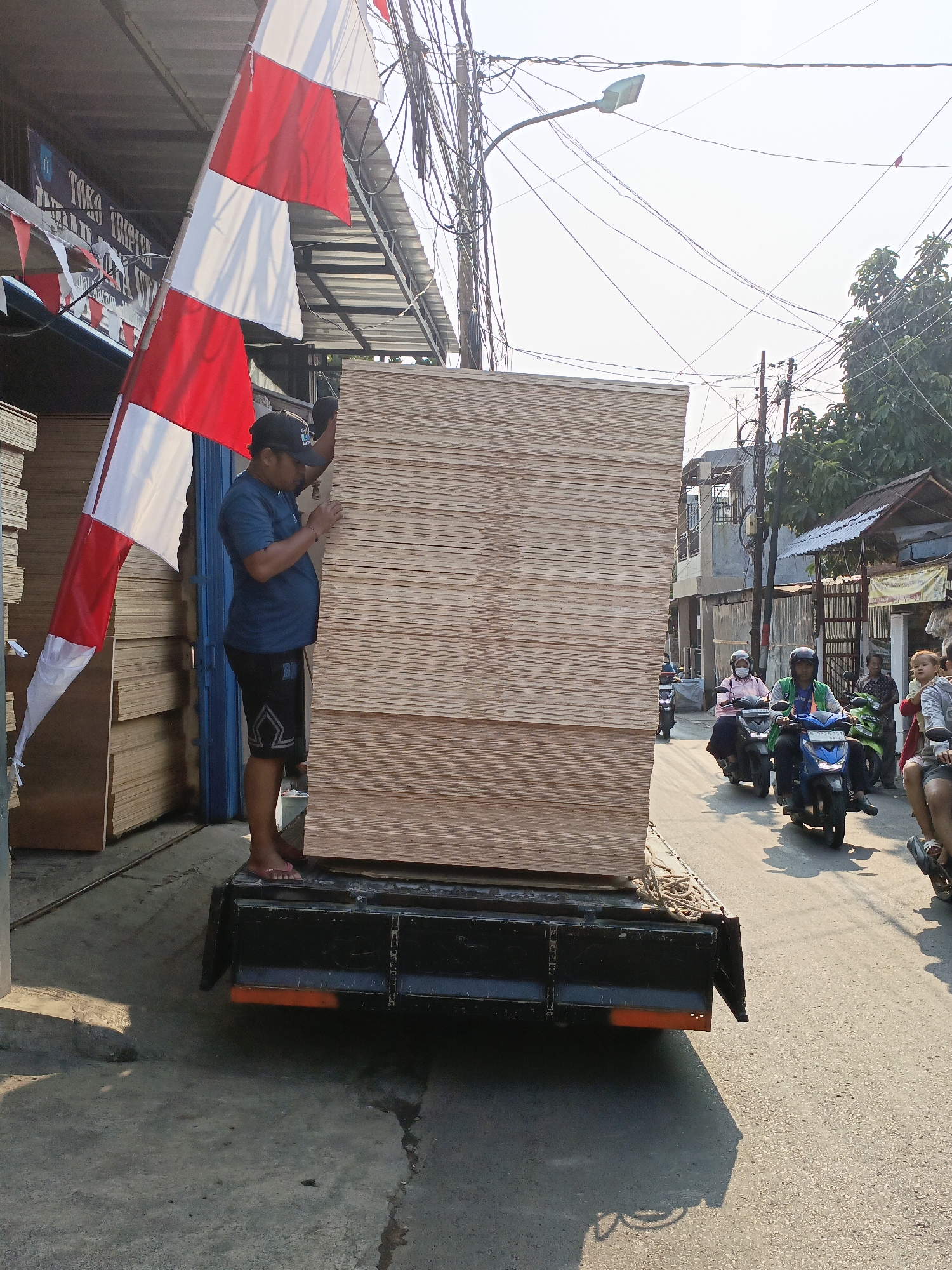 In East Jakarta, Indonesia, a man wearing a blue t-shirt and black shorts stands on the back of a flatbed truck, pushing a large stack of plywood sheets. The plywood is neatly stacked and takes up most of the truck bed. Next to the truck, a tall, striped Indonesian flag (red and white) flutters. The truck is parked on a street with other vehicles, including several motorcycles carrying passengers, in the background. Buildings and utility poles with numerous wires are visible along the street, indicating an urban setting. The scene appears to be a business operating from a roadside location, likely a lumber yard or construction supply store.