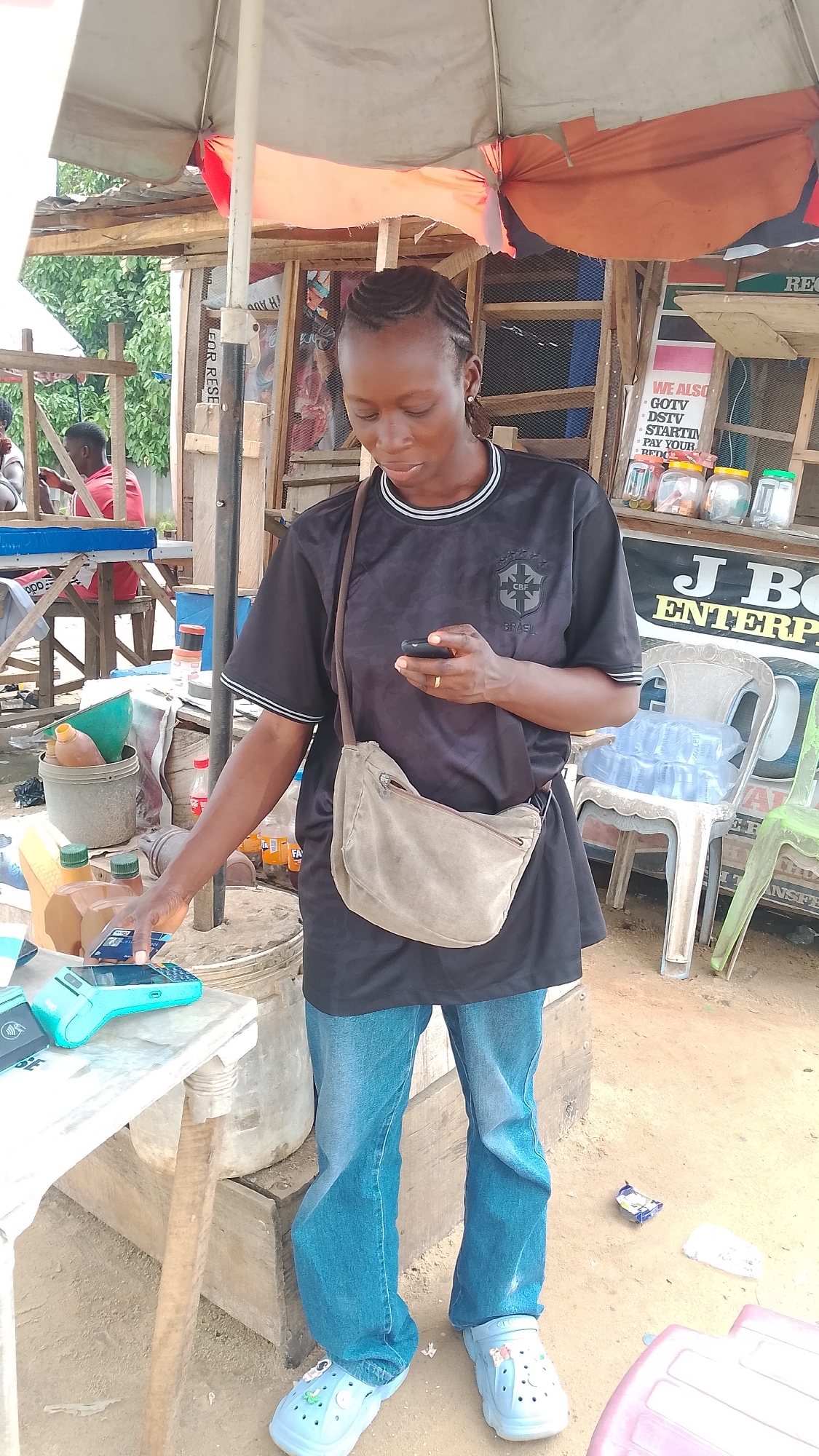 In Warri, Nigeria, a woman with braided hair stands at a market stall, holding a smartphone and interacting with a point-of-sale device. She wears a black t-shirt with a crest and blue jeans, and a light-colored crossbody bag is slung over her shoulder. Her feet are clad in light blue clogs adorned with charms. The stall features a wooden structure covered by an umbrella, with various goods including plastic containers, bottles, and a sign for "J BO ENTERPRISE." In the background, other market stalls and people are visible. The ground is sandy and uneven, typical of an outdoor market.