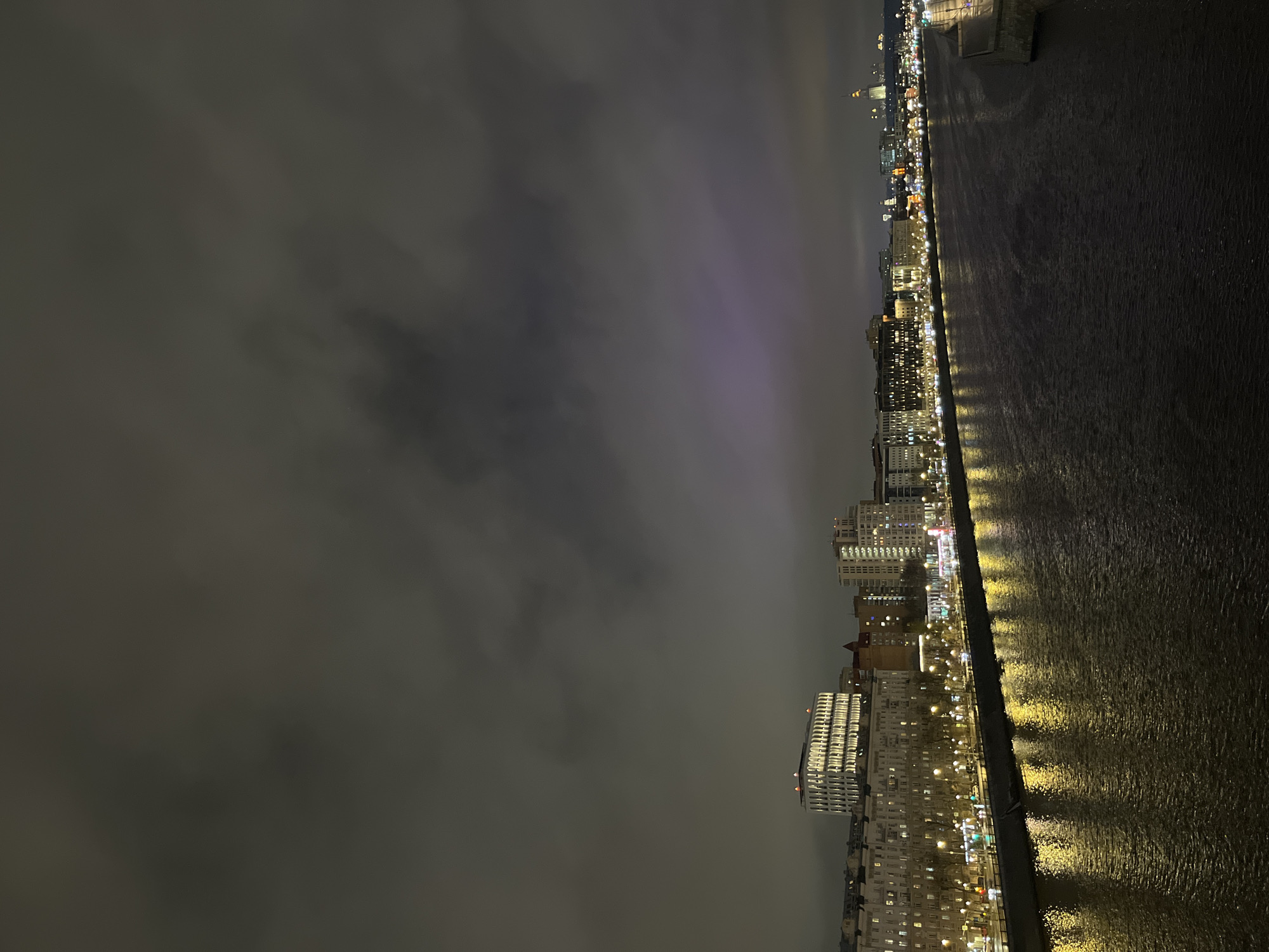 This image shows a nighttime view of the Moscow cityscape along the Moscow River. The sky is dark and overcast. The river reflects the lights from the buildings and streetlights, creating a shimmering effect. Buildings of varying heights are visible along the embankment, with illuminated windows and architectural details. The overall impression is a quiet, urban scene under a cloudy night sky.