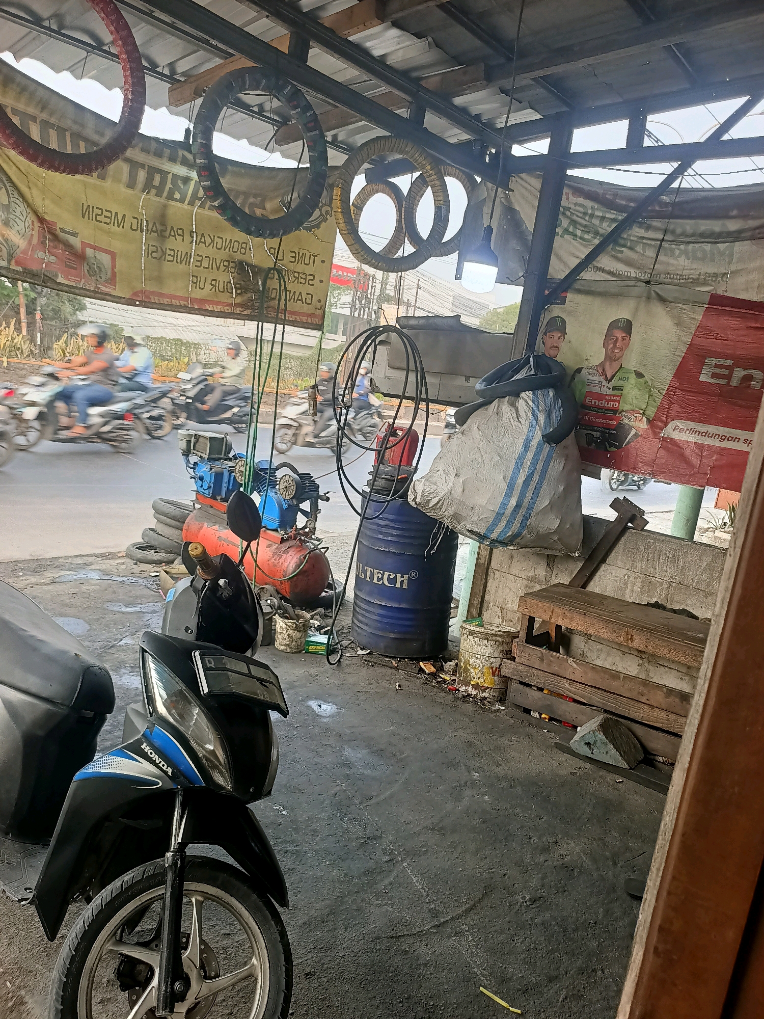 The image depicts a motorcycle repair shop in Gandasari, Indonesia. The shop appears to be open to the street, with several motorcycles visible in motion on the road in the background. In the foreground, a black and blue Honda motorcycle is parked. The shop is cluttered with various tools and equipment, including tires hanging from the ceiling, an air compressor, and barrels. A large banner advertising motorcycle services is displayed prominently. Two men, possibly mechanics or customers, are featured on a poster on the wall. The environment suggests a busy, working-class area.