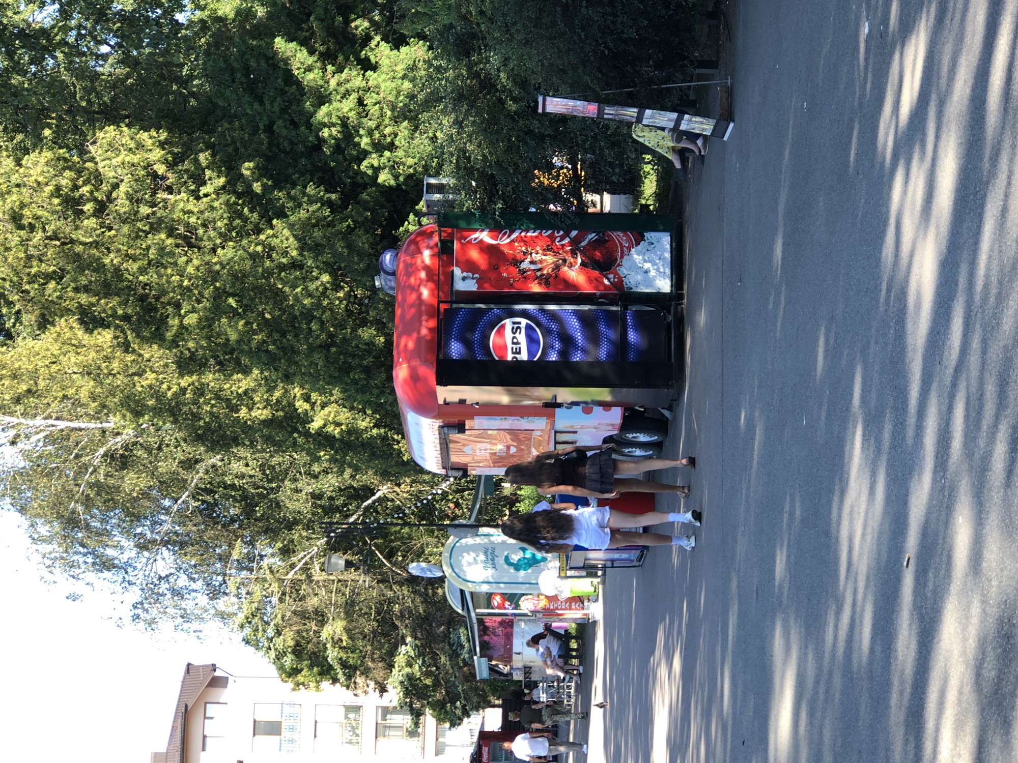 In Kolomyia, Ukraine, a street scene unfolds under a bright sky and dappled sunlight filtering through lush green trees. The foreground is a paved walkway, casting long shadows. To the right, a red and silver food cart stands prominent, its side adorned with a Pepsi advertisement featuring a stylized bottle and icy accents. Next to it, a woman in a yellow floral dress sits on a bench, her legs crossed.
Further left, two young women walk away from the camera, one in white shorts and a white top, the other in a dark top and a dark skirt. Behind them, a cluster of smaller kiosks line the path. One kiosk displays vibrant images, and another has a blue and white design with text. To the far left, a man in a white shirt and dark pants walks past. Beyond the kiosks, a multi-story building with brown trim and balconies is visible. A pigeon walks on the path in the middle ground. The overall impression is a lively, sunlit day in a park or pedestrian area, likely a marketplace or recreational spot.