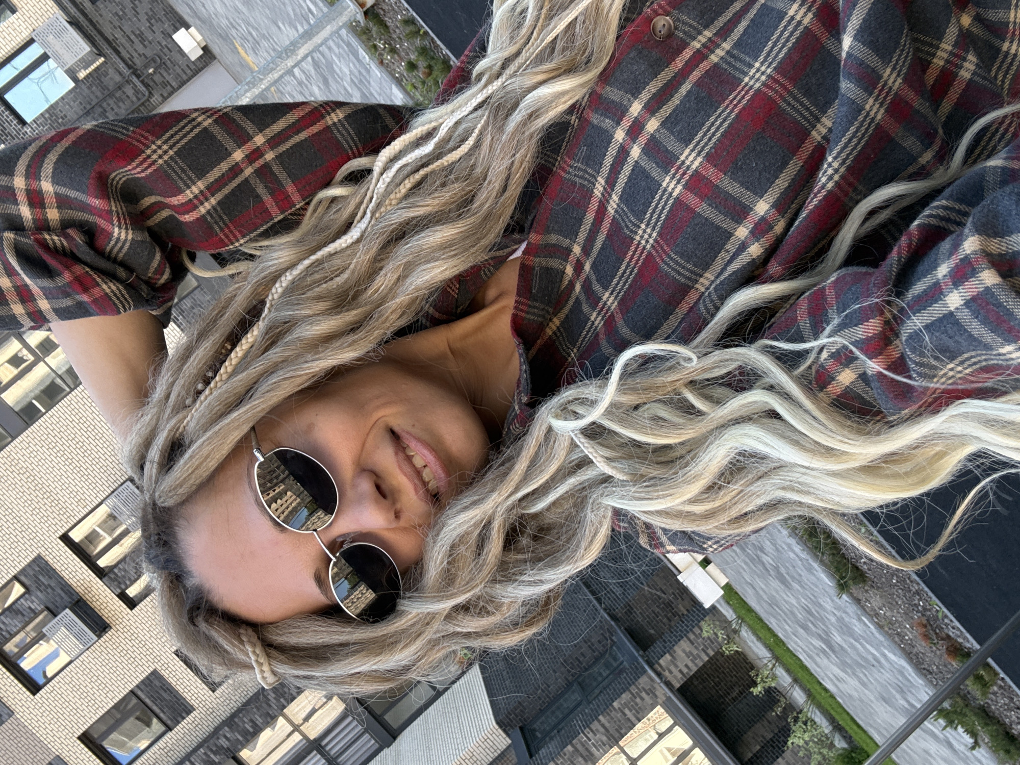 The image is oriented 90 degrees counter-clockwise and features a close-up, high-angle shot of a person against an urban background. The subject is a woman with long, wavy hair that is primarily blonde and light brown, with noticeable lighter braided sections resembling dreadlocks. She is wearing a plaid long-sleeved shirt with a pattern of dark grey, red, and beige squares, and round, dark-lensed sunglasses.
The woman is looking upwards towards the camera with a pleasant smile, revealing a hint of her teeth. Her sunglasses reflect the surrounding environment, including building structures and a bright sky, indicating daytime. One button of her shirt is visible near her neck.
The background, out of focus, shows modern buildings with distinct architectural features such as light-colored brickwork and dark panels. Multiple windows are visible, some reflecting the sky. Below the buildings, there's a suggestion of outdoor space with paved areas and some greenery, possibly a street or courtyard. The overall impression is of a relaxed moment captured in an urban or high-rise setting, possibly from a balcony or rooftop. There is no visible text.