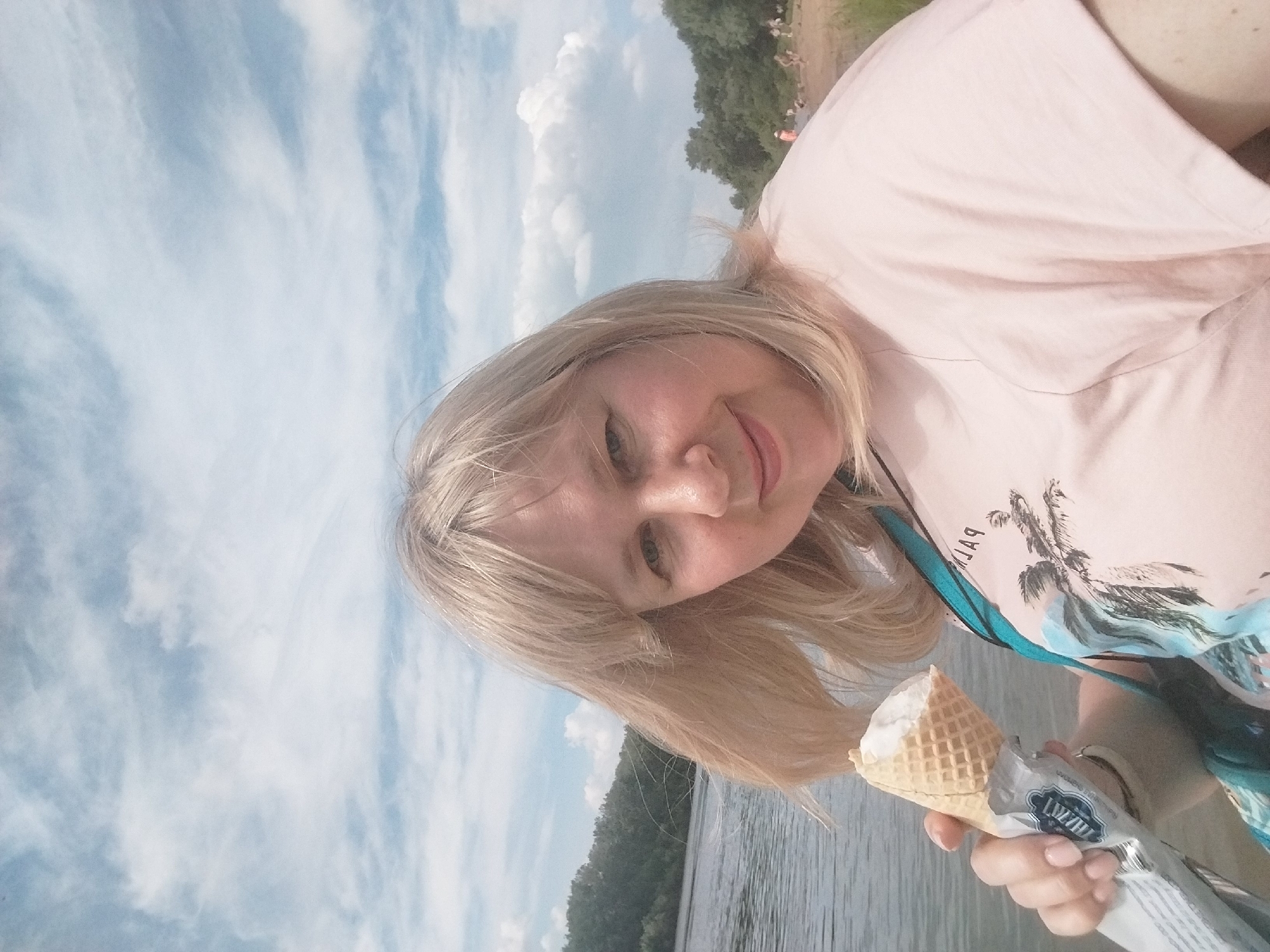 A young woman with blonde hair takes a selfie by a body of water under a cloudy sky. She is wearing a light pink t-shirt with a graphic print of palm trees. She is holding an ice cream cone in her right hand. The ice cream cone has a waffle cone and appears to be vanilla flavored. The background shows a wooded shoreline and a body of water, likely a river or lake. The sky is blue with scattered white clouds. The lighting suggests it is daytime, possibly late afternoon or early evening. The woman has a slight smile on her face. The text visible on the ice cream wrapper is "Пломбир" which translates to "Plombir," a type of Russian ice cream. The overall scene evokes a sense of leisure and enjoyment on a warm day.