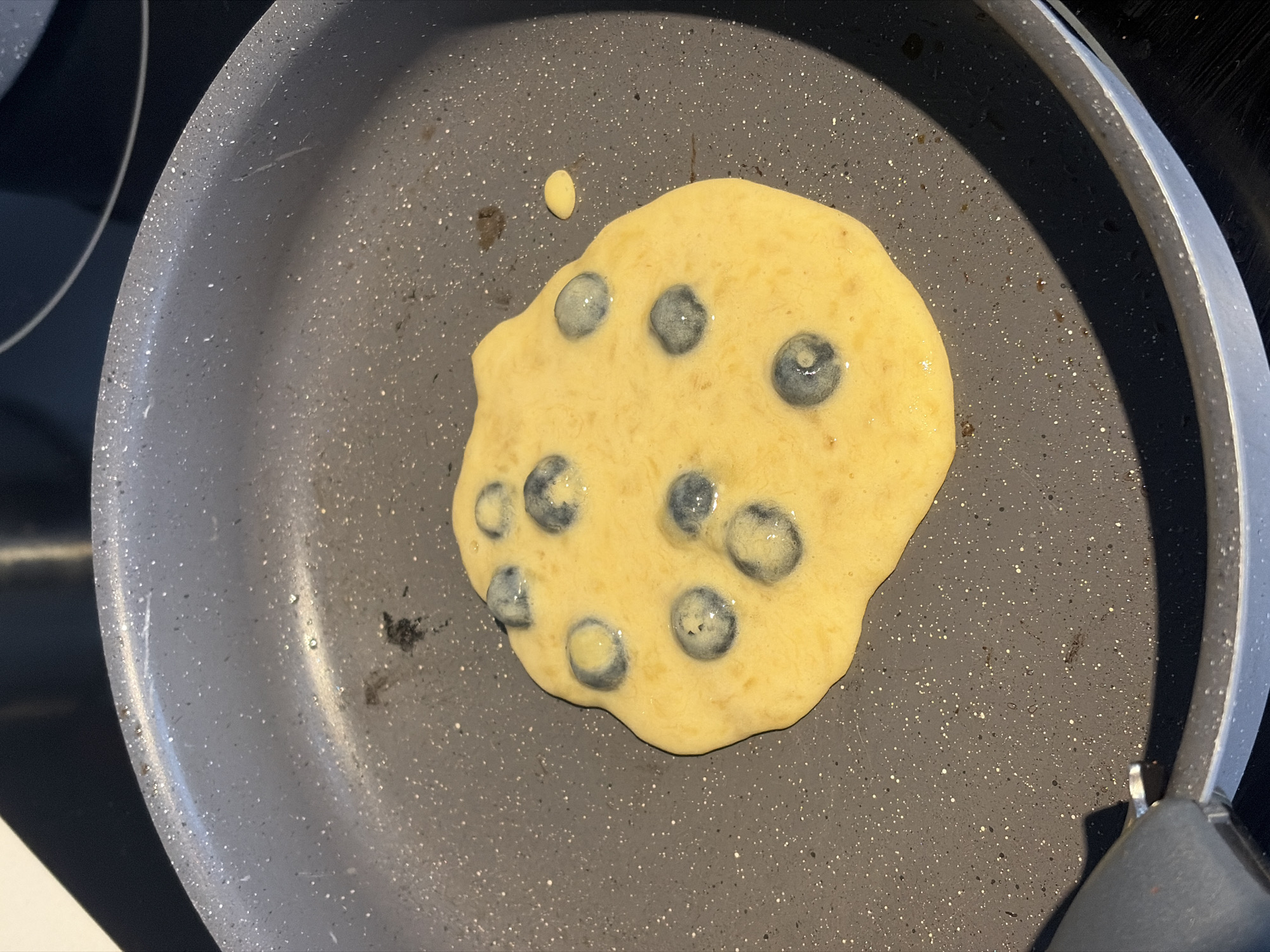 A blueberry pancake cooks in a gray nonstick skillet on a black stovetop. The pancake, slightly off-center in the pan, is a light yellow color and dotted with blueberries. A small droplet of batter sits near the top-left edge of the pancake. The pan's handle is visible on the right side. The location is not discernible from the image. The city of Montreal, Canada, cannot be determined from the image.