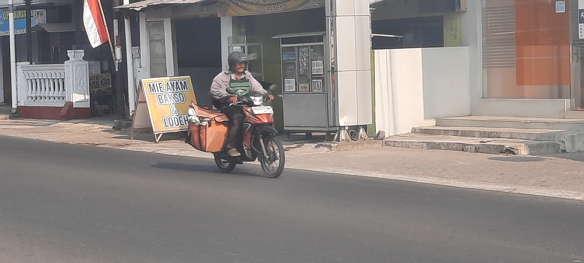 A middle-aged man wearing a helmet rides a red and black motorcycle. He is wearing a grey long-sleeved shirt over a green vest, dark pants, and light-colored shoes. The motorcycle has two orange saddlebags on either side of the rear wheel, filled with white bags. A yellow A-frame sign is visible on the left side of the image, with text in Indonesian. The man is riding on a paved road with a sidewalk to his right. The background shows a building with white pillars, a white railing, and a stall with a grey facade. The location appears to be the city of Selopuro, Indonesia.
