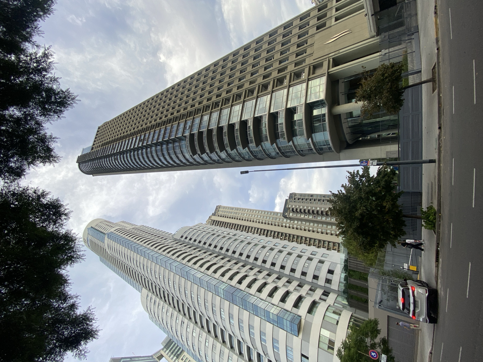 The image shows a street view in Buenos Aires, Argentina, featuring tall buildings framed by trees on the sides. Two distinct architectural styles are evident: one building is a modern, rectangular structure with a lighter facade, and the other is a set of curved, white skyscrapers with blue-tinted glass accents. A street is visible to the right with road markings, a parked silver SUV, and a pedestrian on the sidewalk. The sky is overcast with scattered clouds, suggesting daylight.