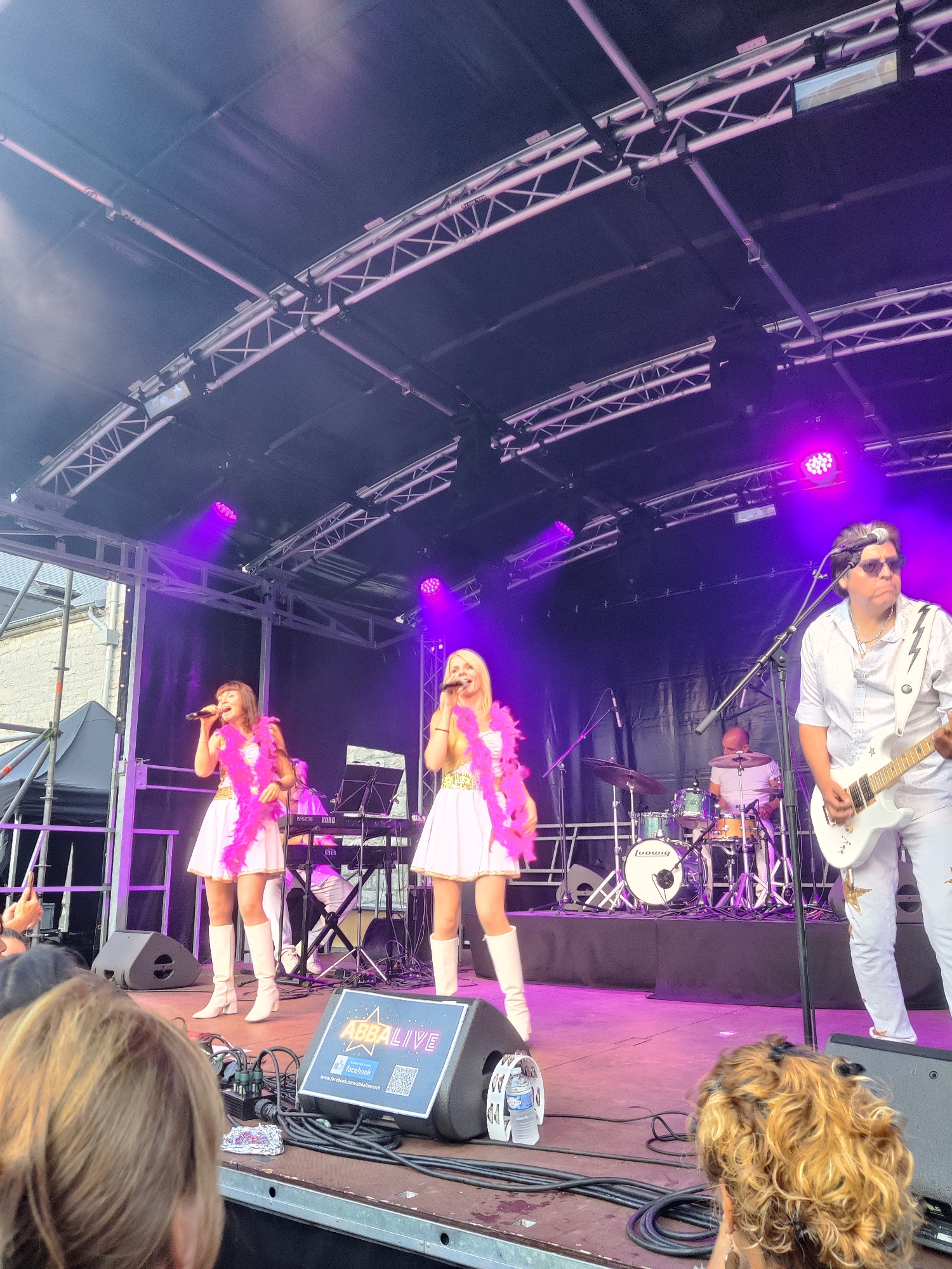 An outdoor concert is in progress on a stage in Marche-en-Famenne, Belgium. The stage is covered by a dark canopy supported by metal trusses, with purple lighting illuminating the scene. On stage, four performers are visible. Two women in the foreground are dressed in white mini-dresses and white boots, adorned with pink feathered boas around their necks. Both are holding microphones and appear to be singing. Behind them, a man in a white shirt and white pants with lightning bolt accents plays a white electric guitar. To the left of the singers, a drum set is positioned, with a drummer partially visible behind it. The stage floor is wooden, and various cables and audio equipment are visible, including speakers. A blue and white sign with "ABBALIVE" and a Facebook logo is prominently displayed on a speaker in the foreground. The audience is partially visible in the lower portion of the image, with their backs to the camera. The background shows a glimpse of a stone building and scaffolding.