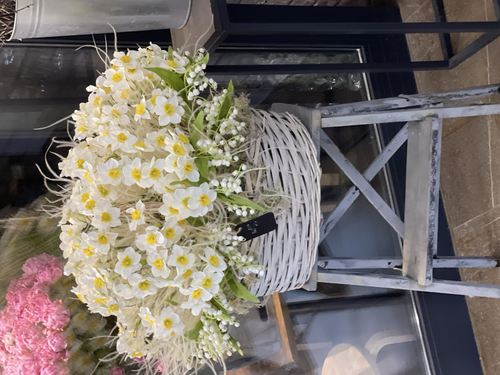 A large basket of white and yellow daffodils and small white flowers is displayed on a rustic wooden step stool. The basket is woven and appears to be made of light-colored wicker. The daffodils are arranged in a dense bouquet, with some greenery and delicate white flowers interspersed throughout. A small black tag hangs from the side of the basket. In the background, blurred pink flowers are visible, along with a metal bucket and a dark window. The scene is indoors, likely in a flower shop or market, possibly in Moscow, Russia.