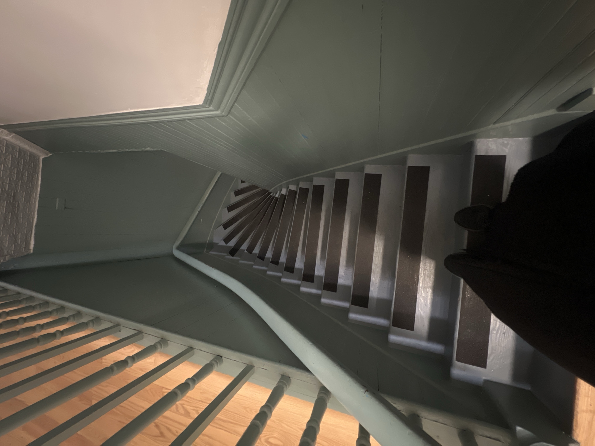 The image shows a downward view of a winding staircase in an interior space. The walls and ceiling are painted in a muted green tone, with white trim. The stairs alternate between a lighter color, possibly gray or white, and a darker, rectangular strip on each step. A black object, likely a person's clothing, is visible on the right side of the frame, partially obscuring the stairs. A wooden railing is present on the left side of the stairs, with wooden bars. The flooring visible through the railing is light-colored wood. There is no indication in the image that places the location in New York City or anywhere else.