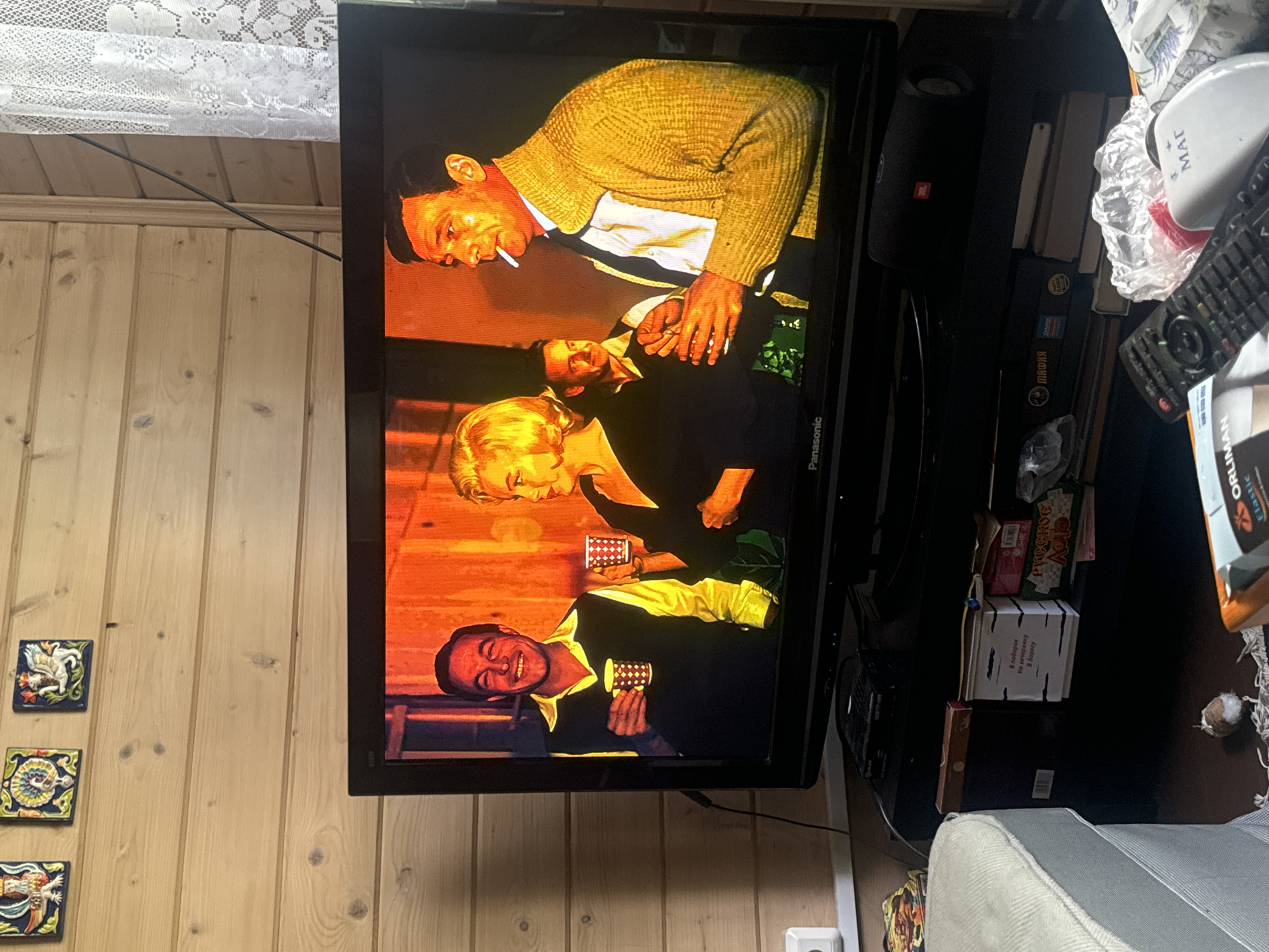 The image shows a television screen displaying a scene from a film or TV show. Three people are visible on screen. A man in the foreground, wearing a yellow sweater over a white shirt and dark tie, is laughing and holding a small checkered cup. Behind him and to the left, a woman with blonde hair, wearing a dark dress, is also holding a similar cup. To the far left, another man is seen wearing a patterned yellow cardigan and smoking a cigarette. The scene appears to be set indoors, possibly in a bar or a dimly lit room, with warm lighting.
To the left of the television, a wooden wall is visible, adorned with three small decorative tiles featuring mythological creatures or figures. To the right of the television, various objects are present on a shelf, including a black speaker with a red JBL logo, a white container with Russian text that appears to read "МАГ" (MAG), a remote control, a white plastic bag, and some boxes. One box has Russian text on it, which is partially legible and reads "РУССКОЕ АЛОЕ" (RUSSKOYE ALOYE) which translates to "Russian Aloe". Another white box with Russian text is also visible, with the text "В секрет. На вечерний. В буфет" (In secret. For the evening. To the buffet).
The overall context suggests a casual setting within a room, with the television as the focal point. The mood on screen appears lighthearted due to the man's laughter. The visible text indicates a Russian context for some of the objects in the scene. The lighting on the television suggests it might be evening or indoors with artificial lighting.