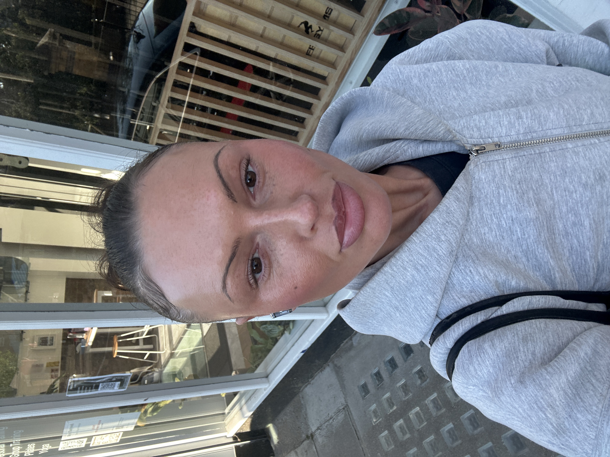 A woman with fair skin, brown eyes, and her hair pulled back is wearing a grey zip-up hoodie with black stripes on the sleeves. She is facing the camera. The background features a storefront window with reflections of buildings and furniture, plus a wooden structure with dark horizontal lines. Outside the building, there is a pavement with square indentations. The scene appears to be in London, United Kingdom, as a sign visible through the window reads "Pilates" and "Yoga".