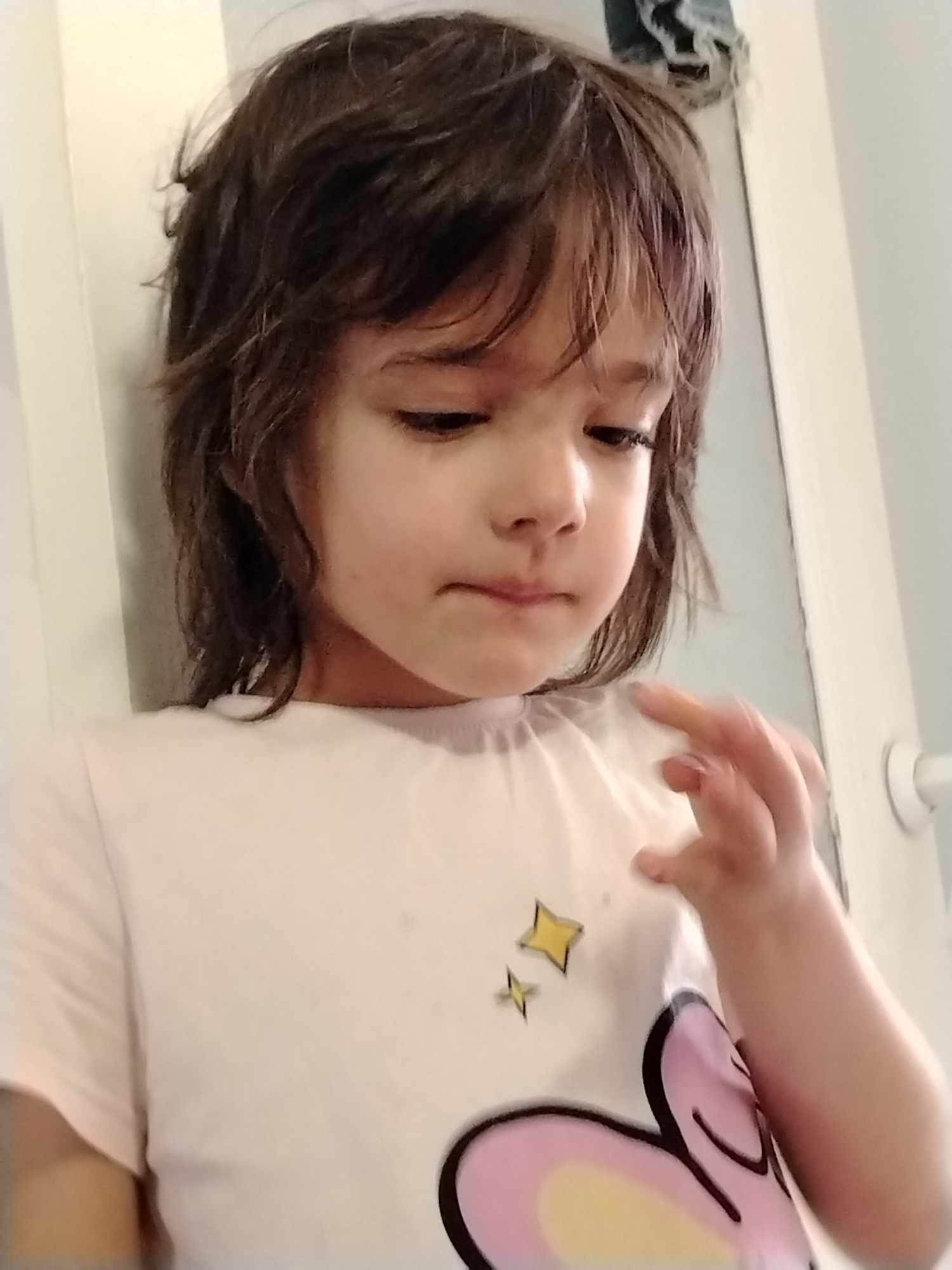 A young child with dark, shaggy hair and brown eyes is looking down in the image. The child is wearing a light-colored t-shirt with a graphic design of a cartoonish animal or character, possibly a bunny, in shades of pink, yellow, and black. Two small yellow stars are also visible on the t-shirt. The child's right hand is raised towards their face, with fingers slightly splayed, and a hint of orange coloring on the index finger, suggesting they might be holding something or have recently touched something colored. The background appears to be indoors, with a white or light-colored wall and a door frame visible on the right. The lighting is soft, casting gentle shadows. There is no visible indication of the location being in Noviy, Russia.