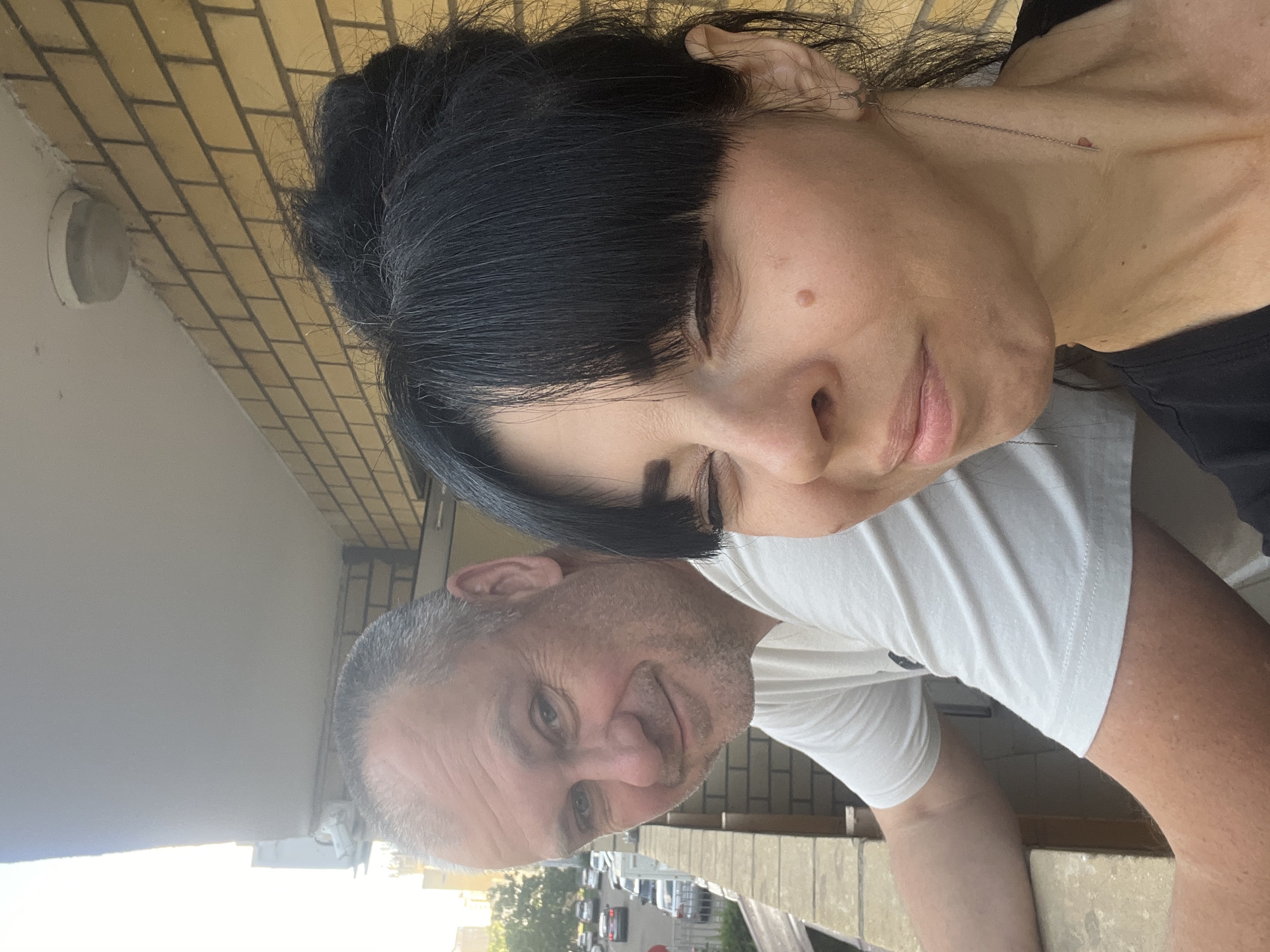 Two people, a man and a woman, are standing on a balcony. The man, on the left, is wearing a white t-shirt and looking towards the camera with a slight smile. He has short, graying hair and stubble. The woman, on the right, is facing the camera and has her eyes partially closed. She has black hair styled in a bun with bangs and is wearing a black shirt. A mole is visible on her right cheek. They are both leaning against a concrete railing. Behind them, a brick wall with yellow bricks is visible, and a light fixture is mounted on the ceiling of the balcony. In the background, cars are driving on a street, and trees and buildings are visible, suggesting an urban environment. The lighting indicates it is daytime. It is not possible to determine the specific location within the city of Krasnodar, Russia from the image alone.