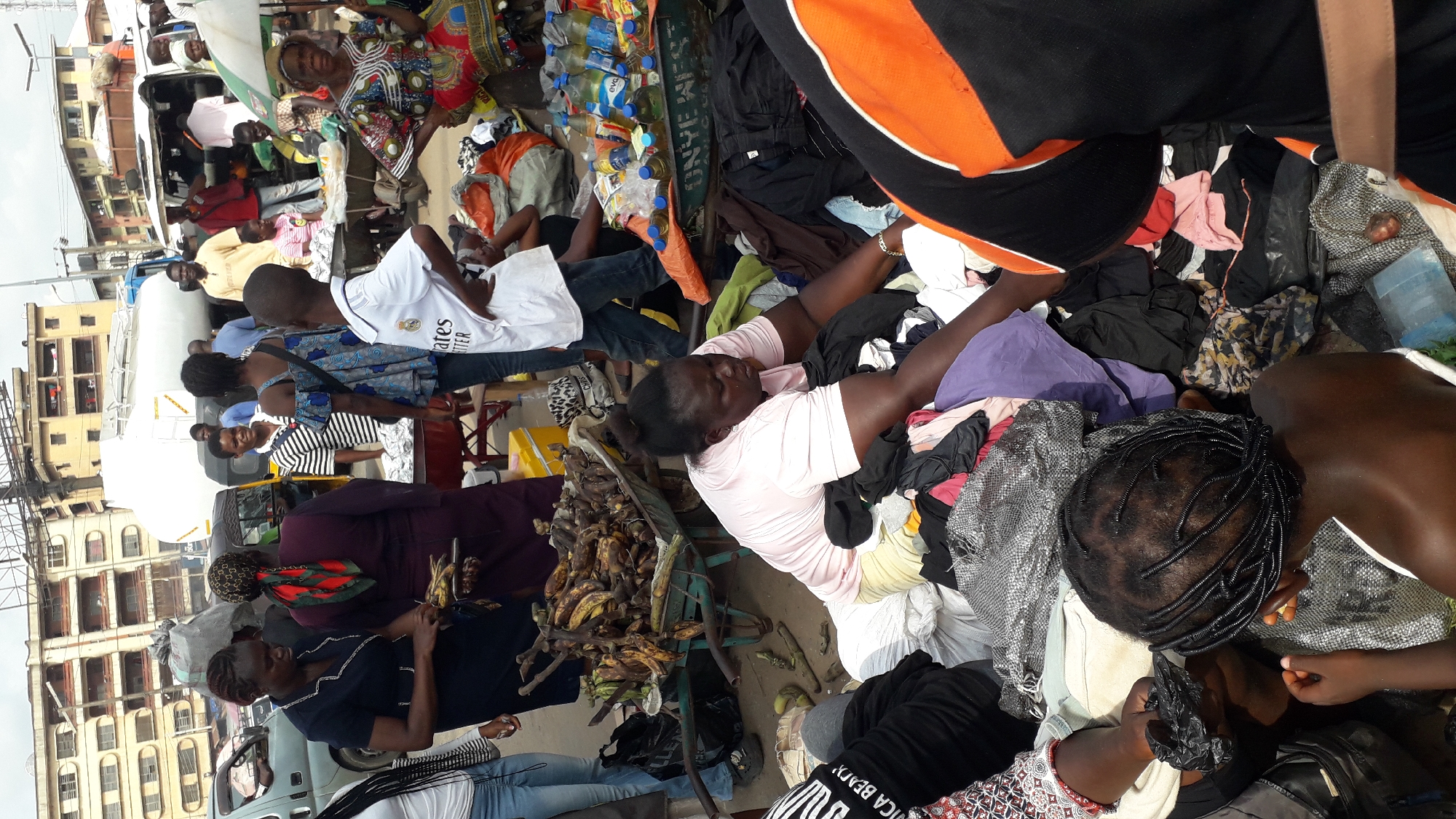 This image depicts a bustling outdoor market scene in Onitsha, Nigeria. The foreground is dominated by a large pile of assorted clothing, with several people interacting with the merchandise. A woman in a pink shirt is sorting through the clothes, while a young child with braided hair sits amongst the garments. In the mid-ground, a wheelbarrow overflowing with bananas is visible, suggesting a produce section of the market. Several individuals are gathered around stalls or carts, some appearing to be vendors or customers. In the background, buildings with multiple stories line the street, characteristic of an urban environment. A large white truck, possibly a water tanker, is also present, along with various other vehicles and people milling about. The overall impression is one of vibrant commercial activity in a densely populated urban setting.