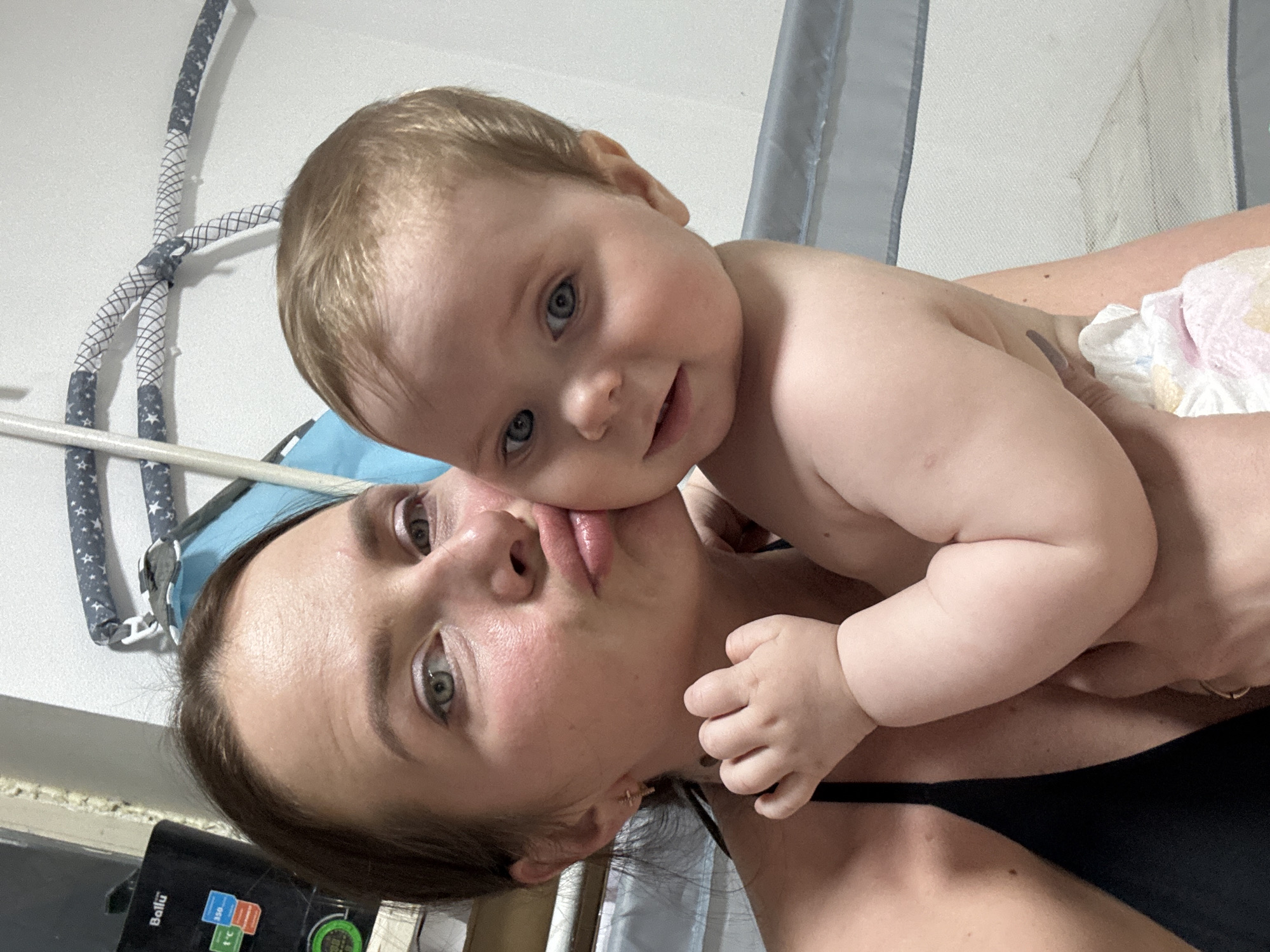 A woman is holding a baby. The woman has fair skin, brown hair, and green eyes. She is wearing a black top. The baby has fair skin, blond hair, and blue eyes. The baby is wearing a diaper. The background is a wall and a crib. There is a Ballu kitchen appliance in the lower left corner. The location appears to be indoors, possibly a home, but the specific city of Rusalevka, Russia is not verifiable from the image.