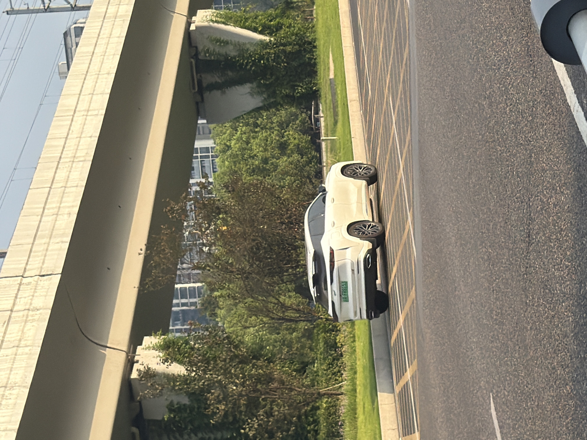 A white SUV is parked on the side of a road under an elevated highway in Jiangjin, China. The SUV has a license plate that reads "A FR955". The road is asphalt and has yellow and white parking lines. There is a sidewalk with a concrete curb separating the road from a grassy area. Trees and buildings are visible in the background. The sky is clear and bright, suggesting it is daytime.