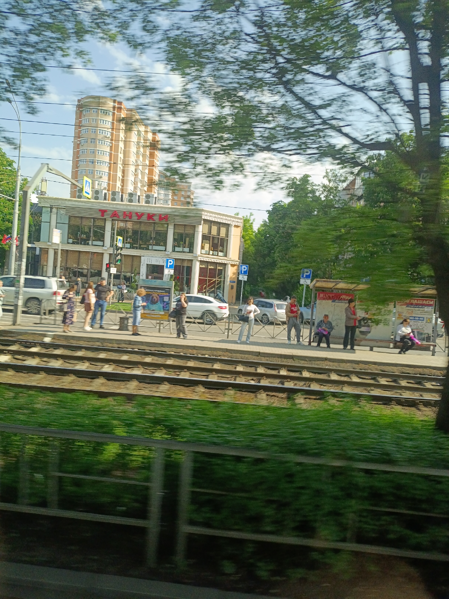 The image depicts a street scene in Krasnodar, Russia, showing a tram stop next to a building with the Cyrillic inscription "ТАНУКИ" (TANUKI), a Japanese restaurant chain. A multi-story residential building is behind it.
Several people are waiting at the tram stop or standing on the sidewalk, some are sitting, and others are standing. There are parked cars visible along the street. A tram track runs parallel to the sidewalk. The image appears to be taken from a moving vehicle, as evidenced by the motion blur on the foliage in the foreground.