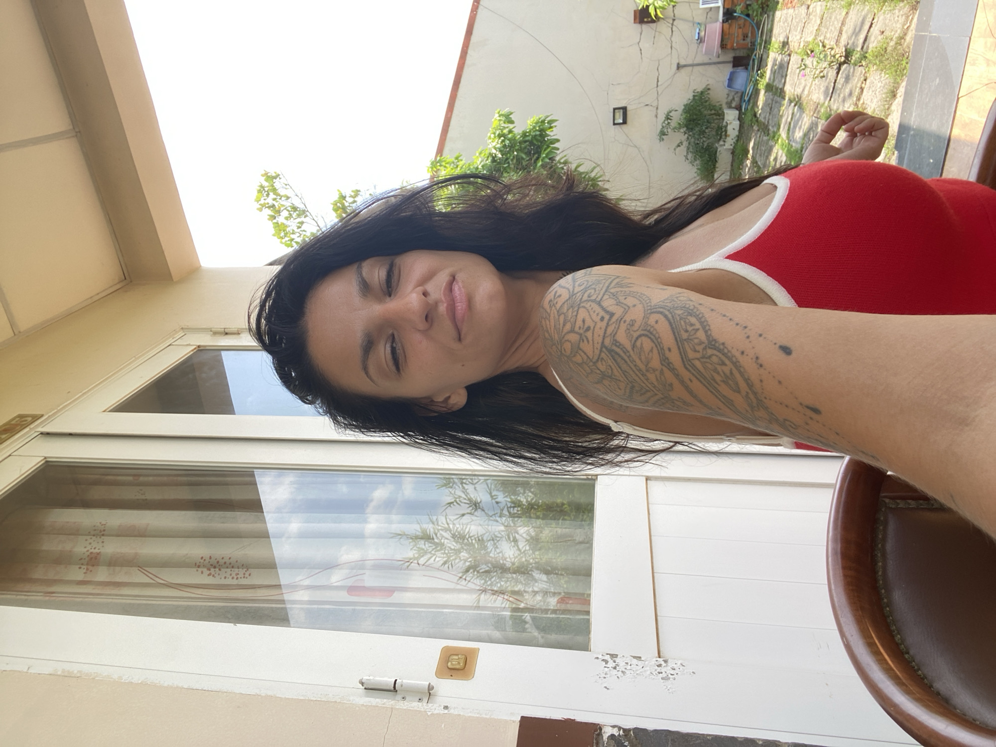 A woman with long, dark hair and a large tattoo on her left arm is taking a selfie outdoors. She is wearing a red tank top with white trim. She is sitting in a brown leather chair, and behind her is a white door with glass panels, revealing vertical blinds and reflections of trees and the sky. To the right of the door, a white wall is visible, with some greenery and outdoor equipment on the ground. The setting appears to be a residential area, possibly a house or guesthouse, in Mũi Né, Vietnam, indicated by the architecture and vegetation.