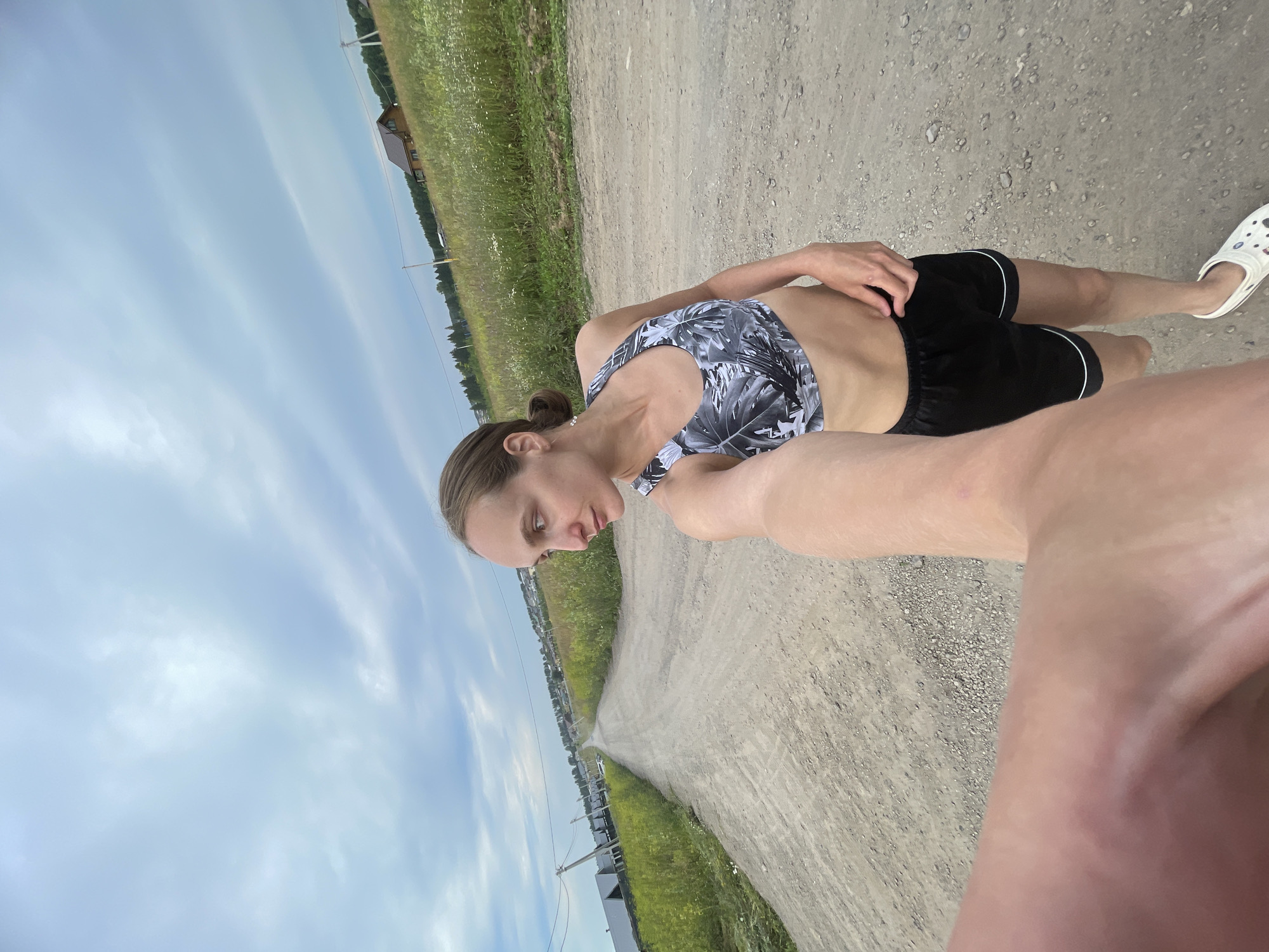 A young woman is standing on a dirt road, looking down and to her left. She is wearing a black and white leaf-patterned sports bra and black shorts. She has her left hand on her hip and her right hand is out of frame, likely holding the camera. She is barefoot on her right foot, wearing a white Croc on her left. The road is surrounded by green grass and fields. In the background, there are a few houses and trees, suggesting a rural or suburban setting. The sky is partly cloudy and a soft blue. The lighting suggests it is daytime, likely late afternoon or early evening due to the soft light. The woman's expression is pensive. The image is taken from a selfie perspective. The overall mood is peaceful and relaxed. The location is Podshivalovo, Russia. There is no visible text in the image.