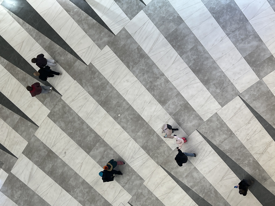 The photograph shows an aerial view of a patterned tile floor with people walking on it. The floor has a linear design with alternating gray and white stripes creating a geometric pattern. Six clusters of individuals are present, all appearing small due to the perspective. One person stands apart from the others. The style of the photo seems candid, emphasizing the floor's design and the movement of people through the space.