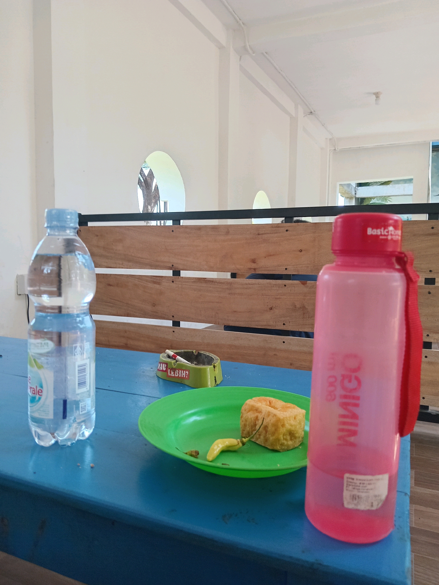 In a casual outdoor dining setting, a blue wooden table is laden with various items. To the left, a clear plastic bottle of mineral water stands, its label partially visible. Next to it, a small green ashtray contains a lit cigarette and is adorned with a sticker that reads "MAU LUGIN?". In the center, a bright green plate holds a piece of fried food, possibly tofu or tempeh, accompanied by a single yellow chili pepper. To the right, a pink plastic water bottle with a red strap is prominently placed. The background features a rustic wooden wall with horizontal planks, and beyond that, a glimpse of an arched opening revealing a tree. The overall scene suggests a moment of relaxation or a break, possibly in Podorejo, Indonesia, given the context.