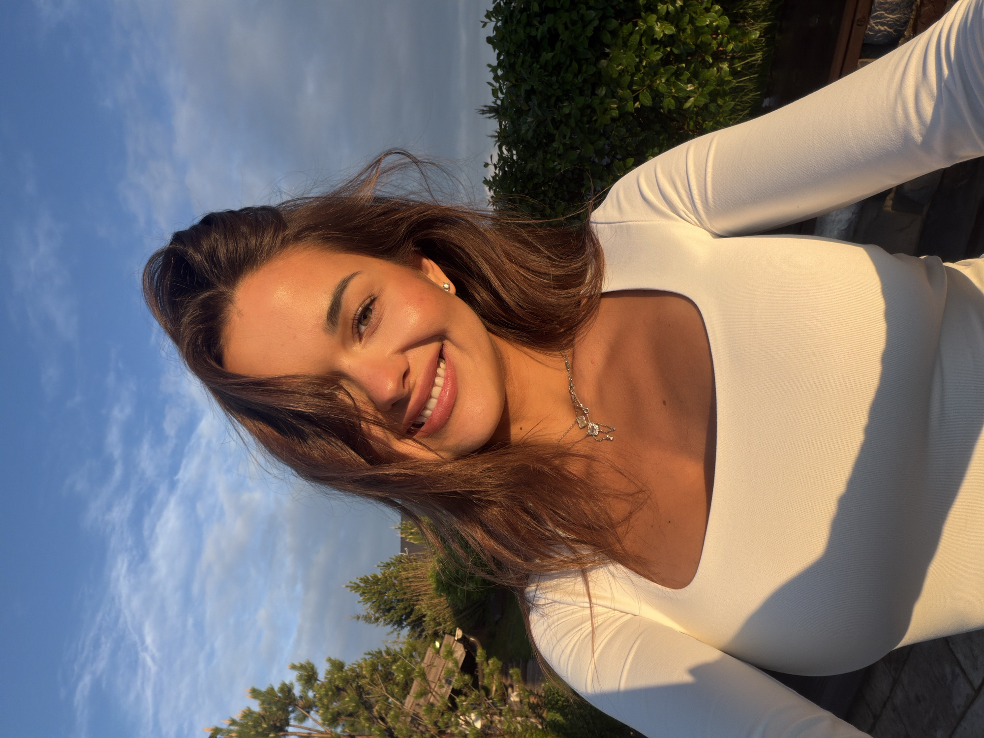 A young woman with brown hair is smiling at the camera. She is wearing a white, long-sleeved top and a delicate silver necklace. The background consists of a blue sky with some clouds, and greenery, possibly trees or bushes. The lighting suggests it's either sunrise or sunset. The photo appears to be taken outdoors, potentially in Элита, Russia, though no specific landmarks are visible to confirm the location definitively.