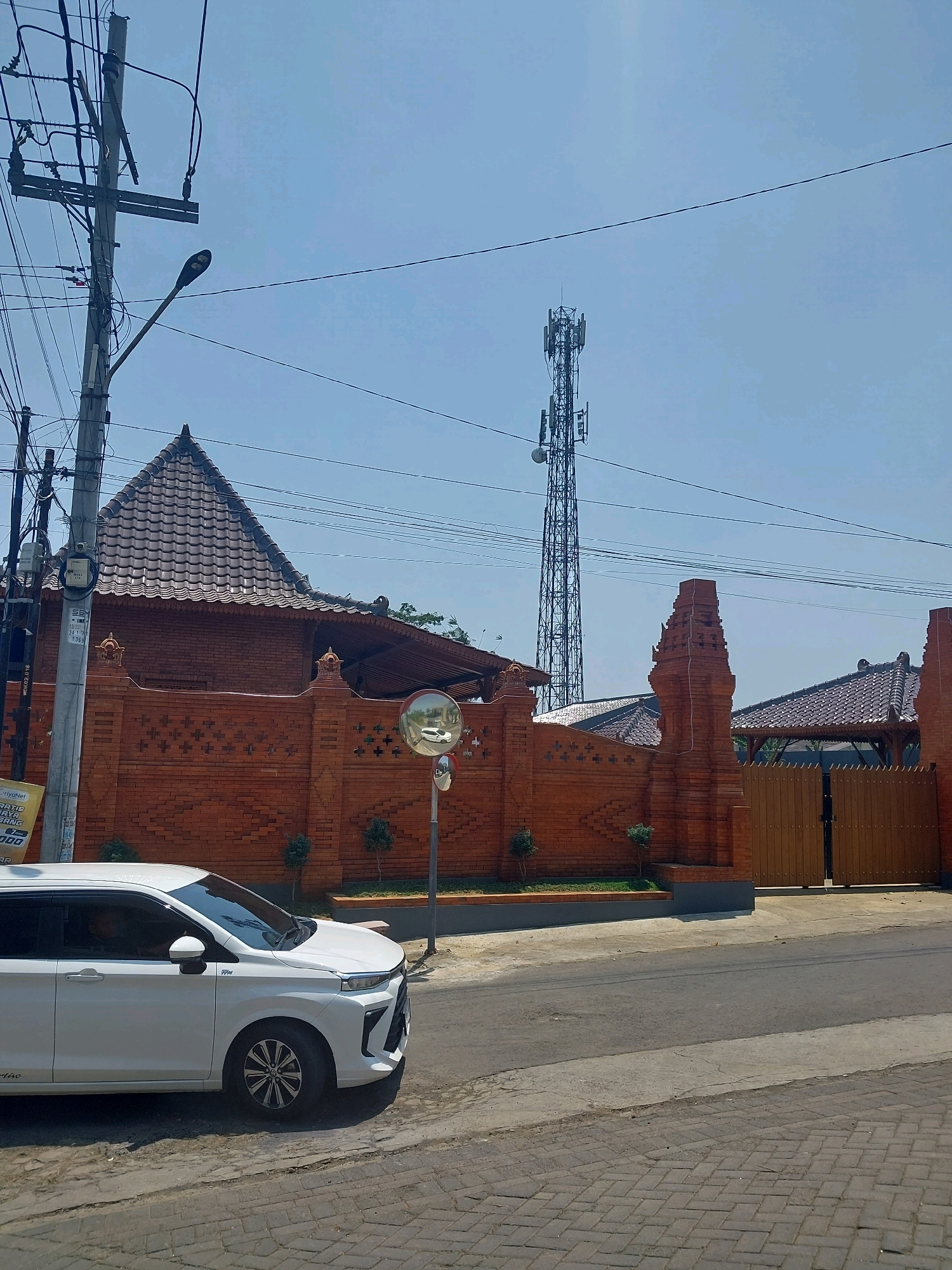 The image depicts a street scene in Malang, Indonesia. A white multi-purpose vehicle is parked on the left side of the road, facing right. Behind the vehicle, a brick wall with decorative elements encloses a property. A traditional Indonesian-style roof is visible above the wall. In the background, a tall telecommunications tower stands against a clear blue sky, with power lines crisscrossing the frame. A street lamp is also present on the left side, attached to a utility pole. The road in the foreground is paved with cobblestones and asphalt. The overall impression is a sunny day in a semi-urban environment.