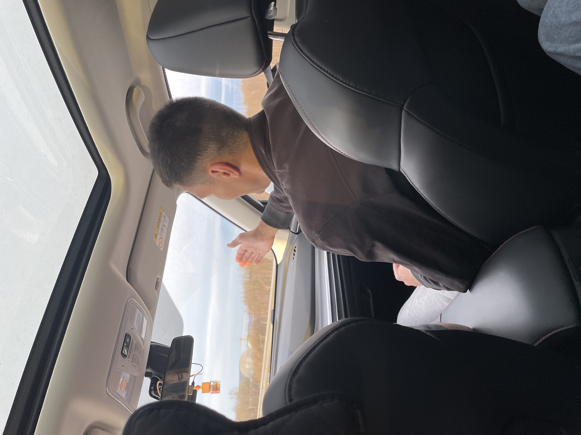 The image captures an interior view of a car, looking forward and to the passenger side, during what appears to be a clear, bright day. A male individual, seen from the back and side of his head, is prominently featured leaning out of the open passenger window, with his right arm and hand extended outward. He is wearing a dark brown jacket.
The car's interior features black leather seats, with subtle red stitching visible on the headrests. The ceiling and pillars are light beige or grey. An overhead console is visible, equipped with various buttons and indicator lights, one of which emits a green glow. A wide sunroof is partially visible, contributing to the bright lighting inside the car. A rear-view mirror is equipped with an attached dashcam or similar device, from which a small, orange rectangular air freshener or charm hangs. An "AIRBAG" warning label is present on the car's A-pillar near the windshield.
Beyond the car window, a bright sky stretches above a landscape populated by what appear to be bare trees lining a road or path, suggesting a rural or semi-rural environment, possibly during late autumn, winter, or early spring.
Parts of two other individuals are also visible: the torso of a person in the front passenger seat, wearing a light grey long-sleeved shirt, and the leg of someone in the back seat behind the leaning individual, wearing grey sweatpants. The scene depicts a passenger interacting with the outside environment from the vehicle. The location context is Kitsjanovo, Russia.